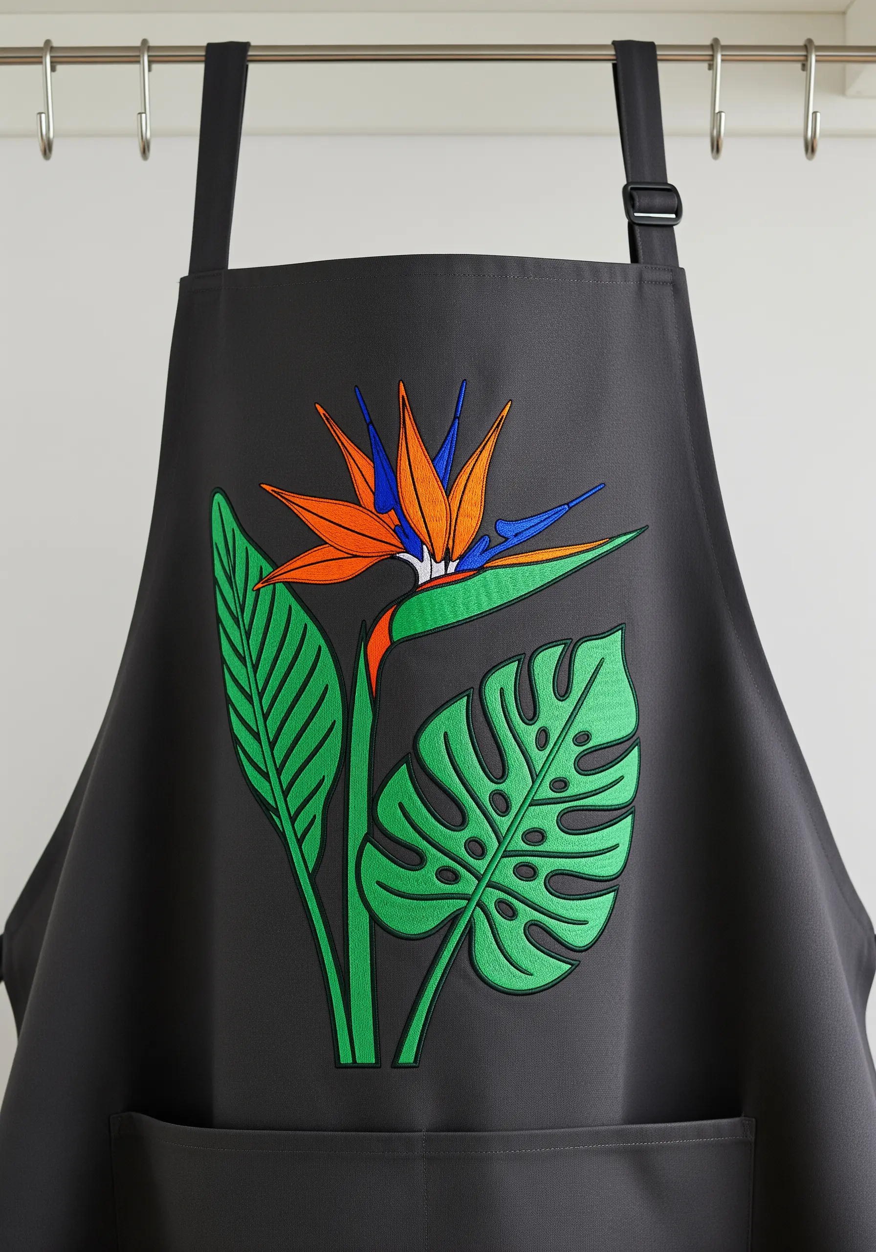 A graphic-style embroidery of a Bird of Paradise and Monstera leaf on a black apron.