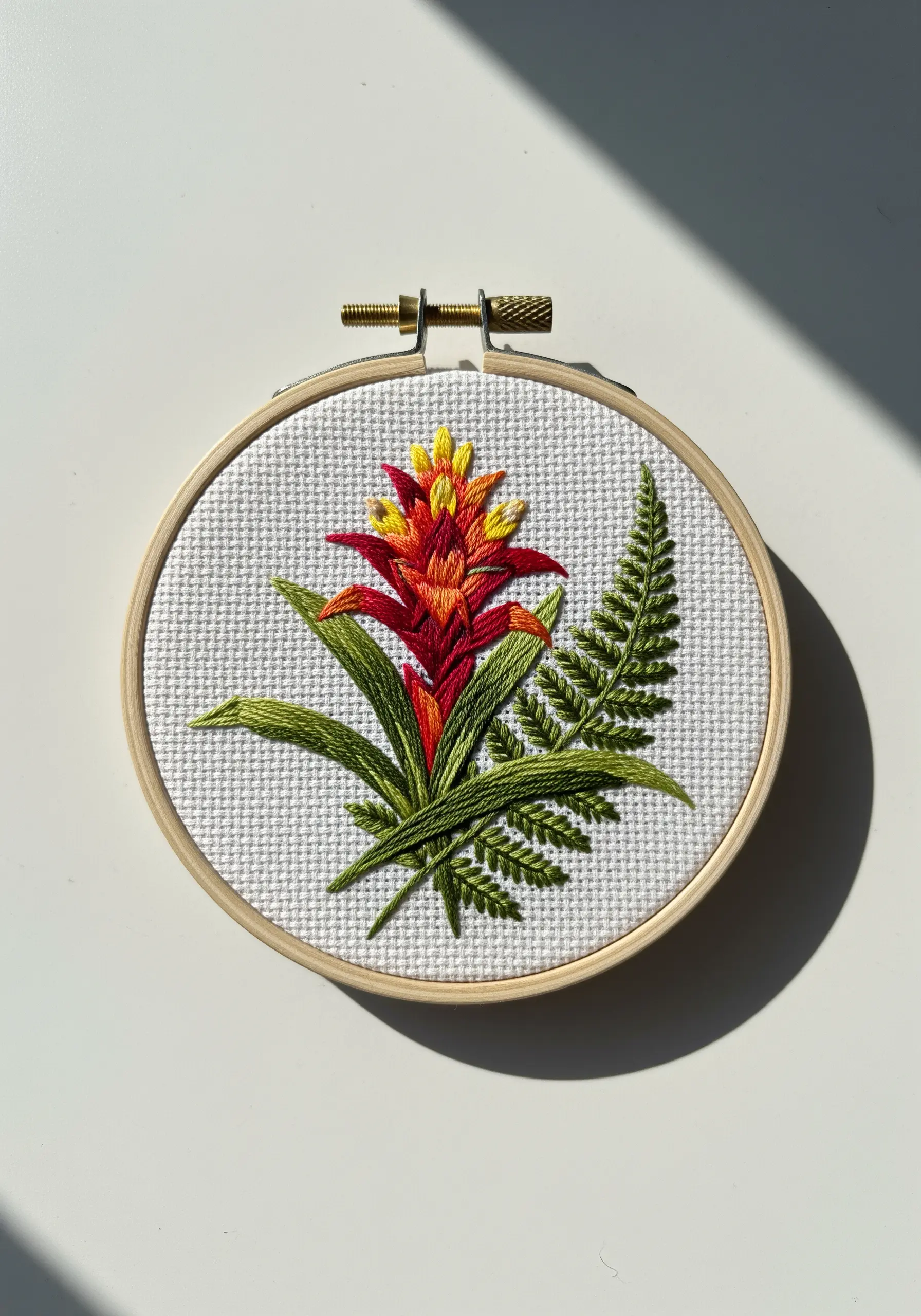 A bromeliad flower and a fern leaf embroidered in a hoop on white Aida cross-stitch fabric.