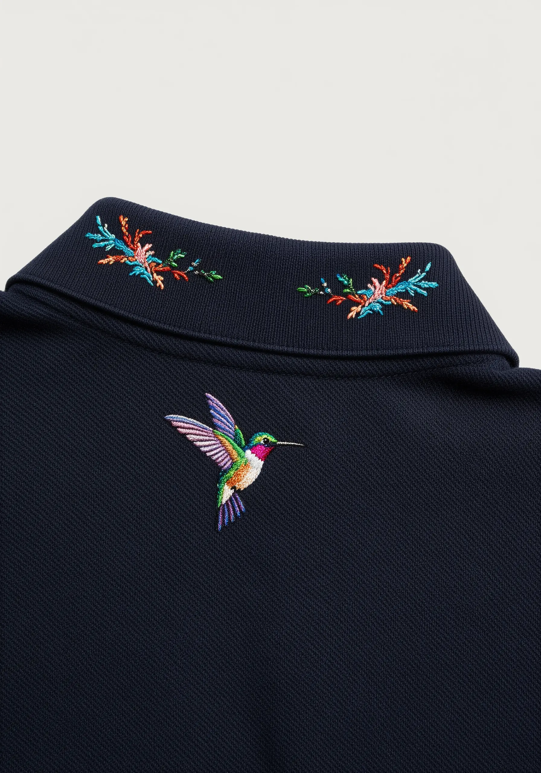 A colorful embroidered hummingbird beneath the back collar of a navy blue polo shirt.