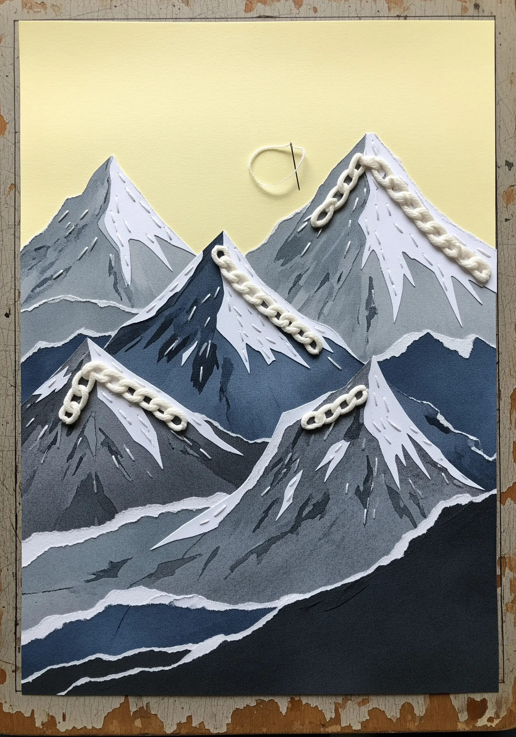 A paper collage of mountains with white chain stitch embroidery depicting snowy paths.
