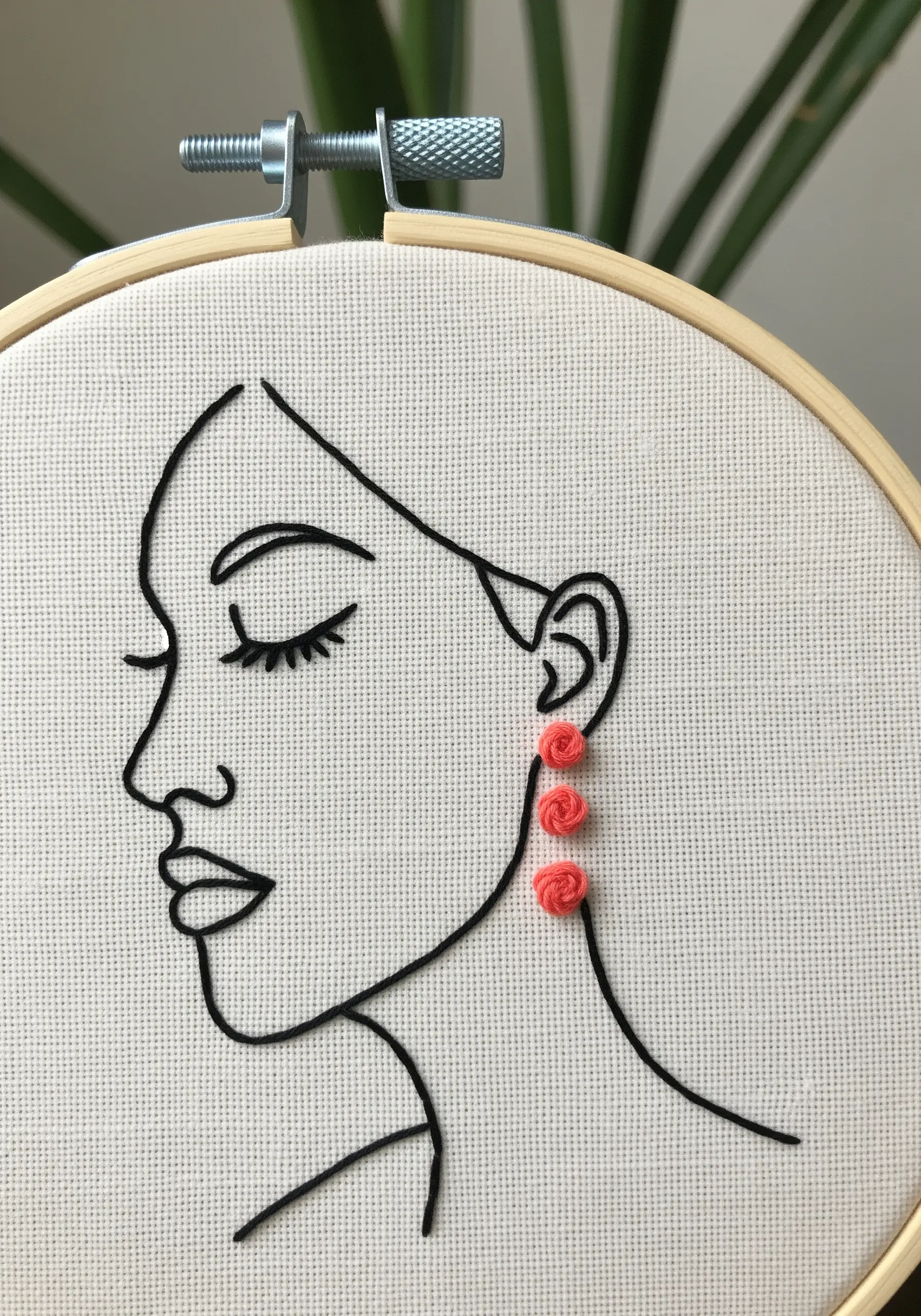 A black line art face in profile, with three tiny, textured coral-pink woven wheel roses as earrings.