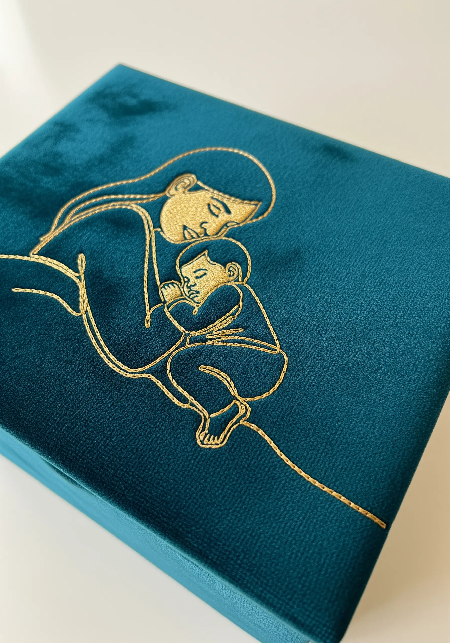 A mother-and-child line art design stitched in gold metallic thread on a teal velvet box lid.
