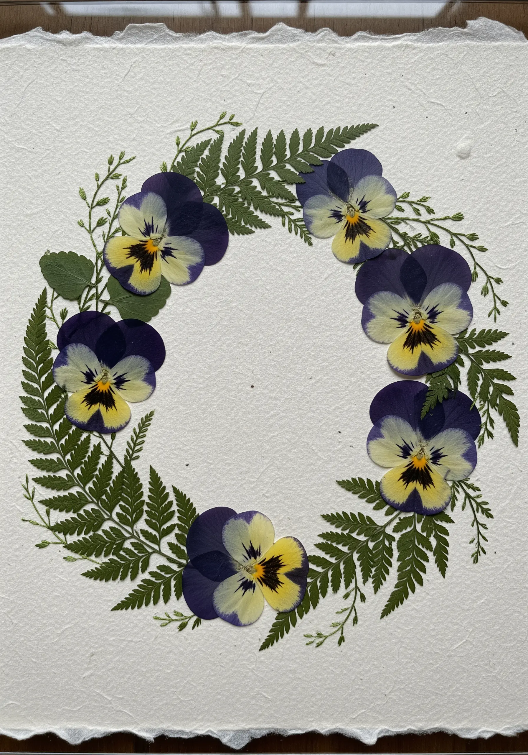 A wreath made of real pressed pansies and ferns arranged on a sheet of textured handmade paper.