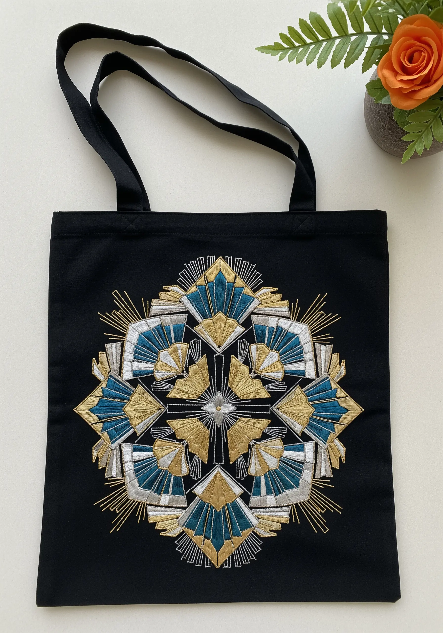 An Art Deco-style geometric mandala in gold, silver, and teal on a black tote bag.