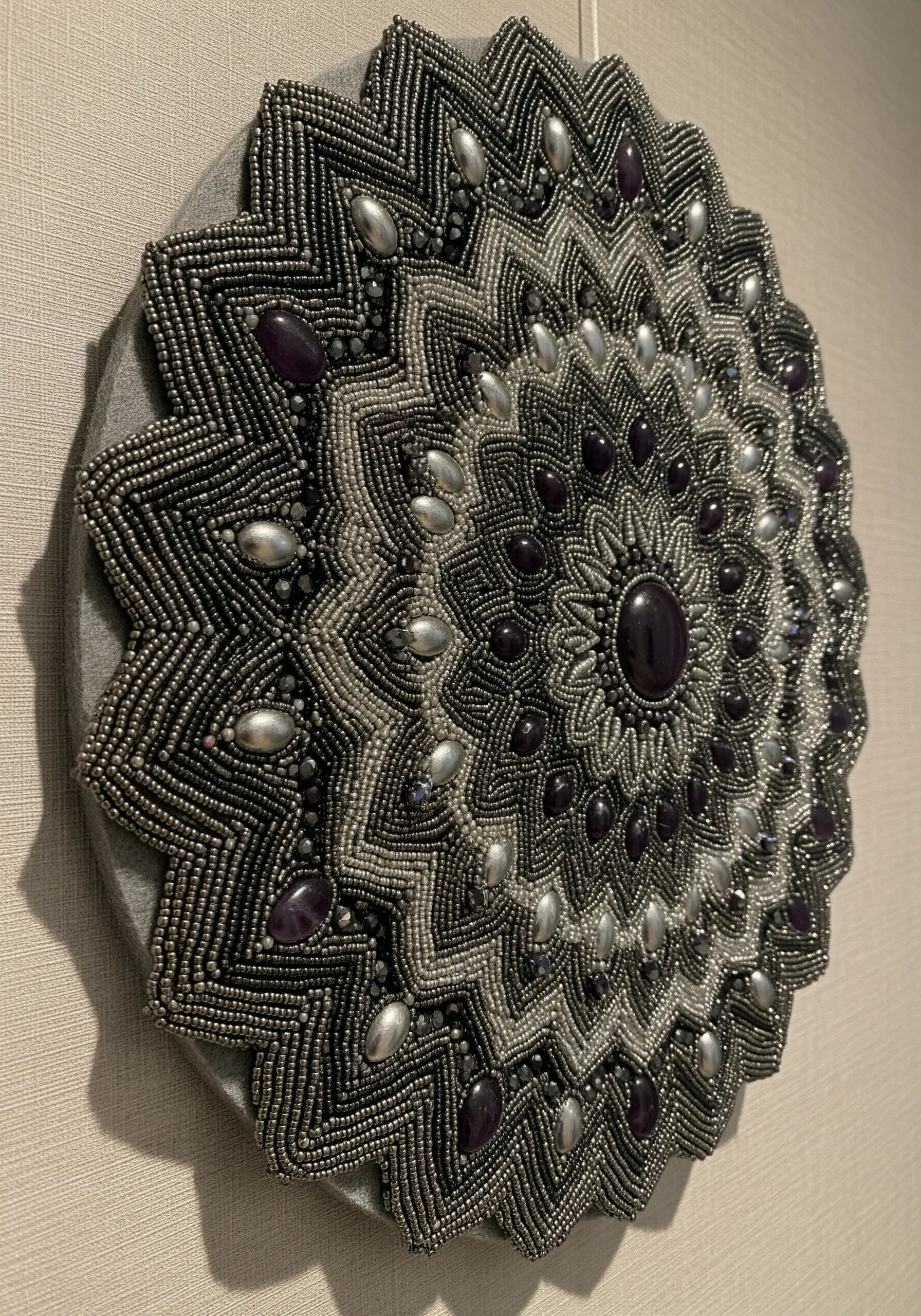 A highly detailed, three-dimensional mandala made entirely of metallic and purple beads on gray felt.