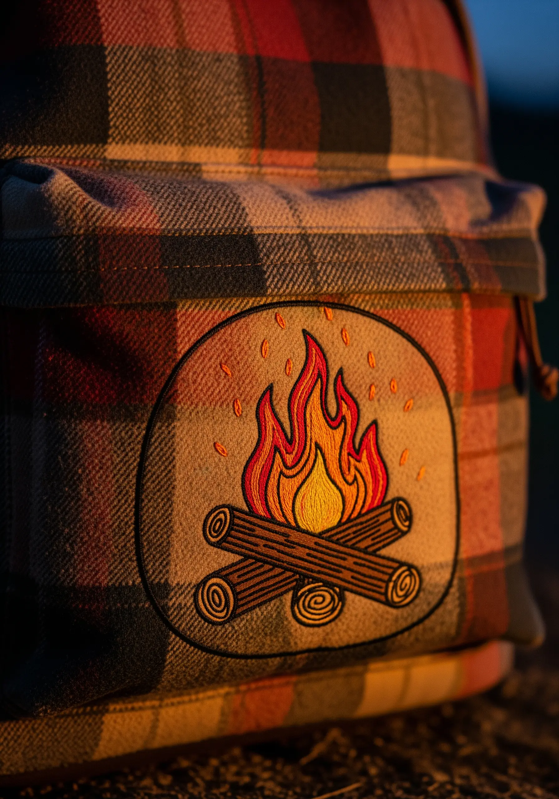 A colorful embroidered patch of a campfire on a plaid flannel backpack.