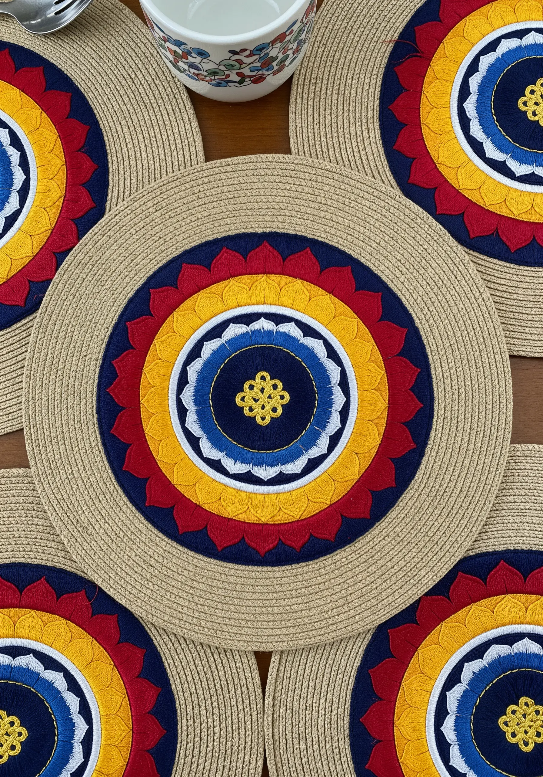 A round placemat with a colorful, embroidered mandala of red, yellow, and blue.