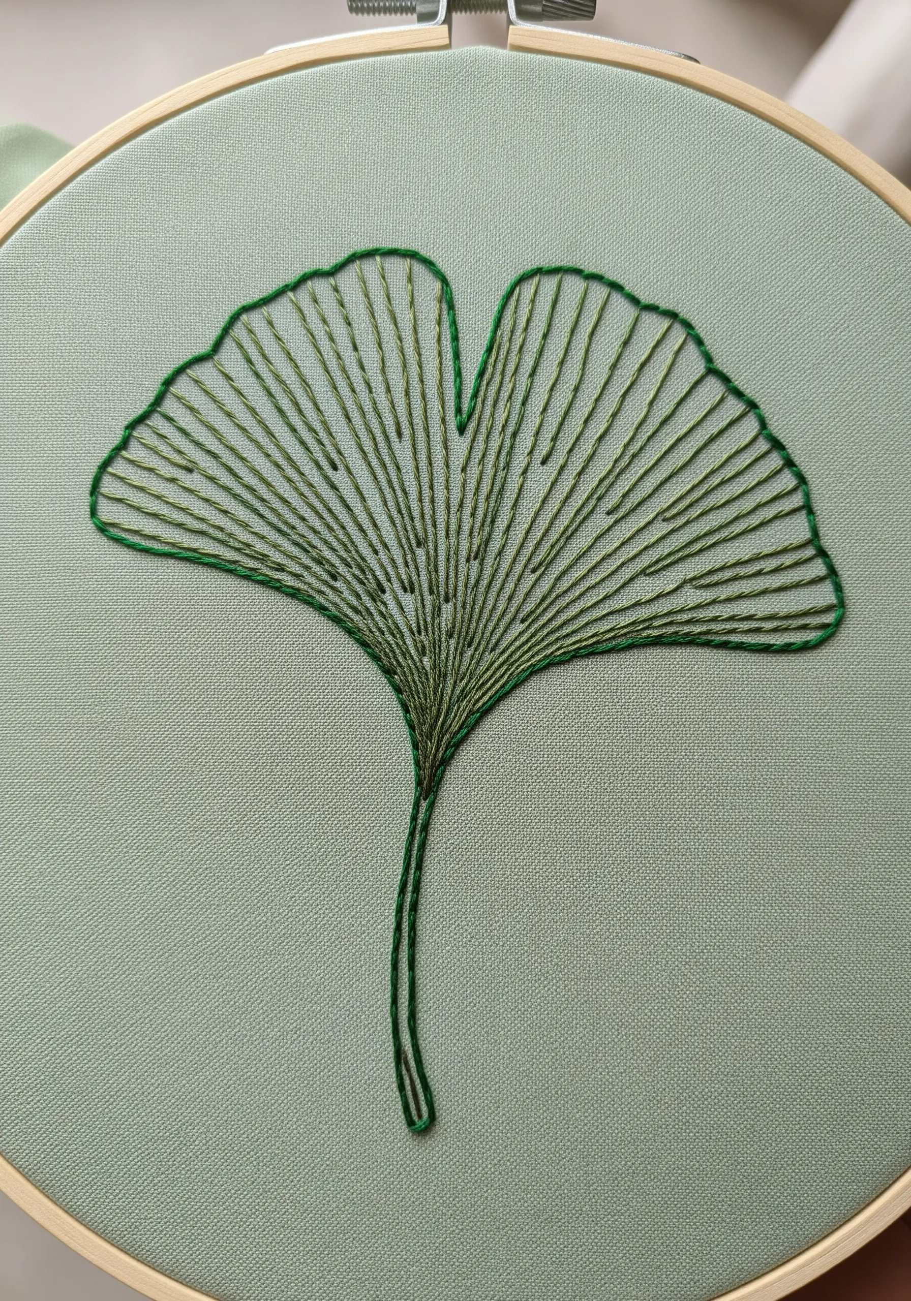 An embroidered ginkgo leaf with radiating straight stitches on a sage green fabric.