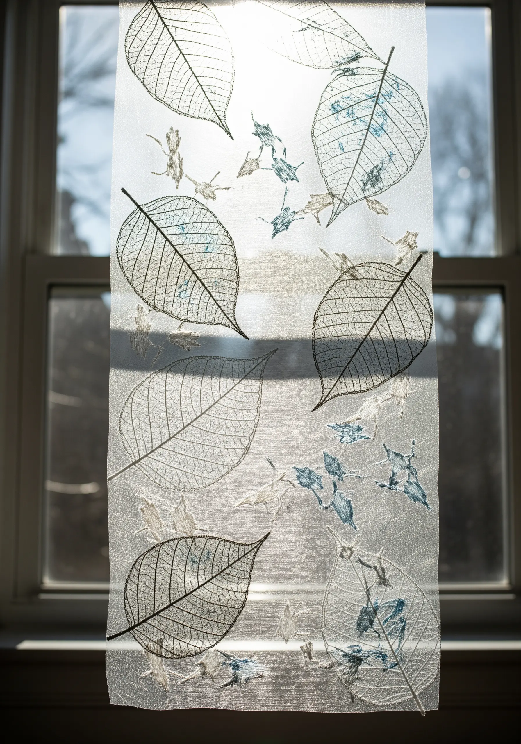 Sheer window hanging with embroidered skeleton leaves, allowing sunlight to shine through.