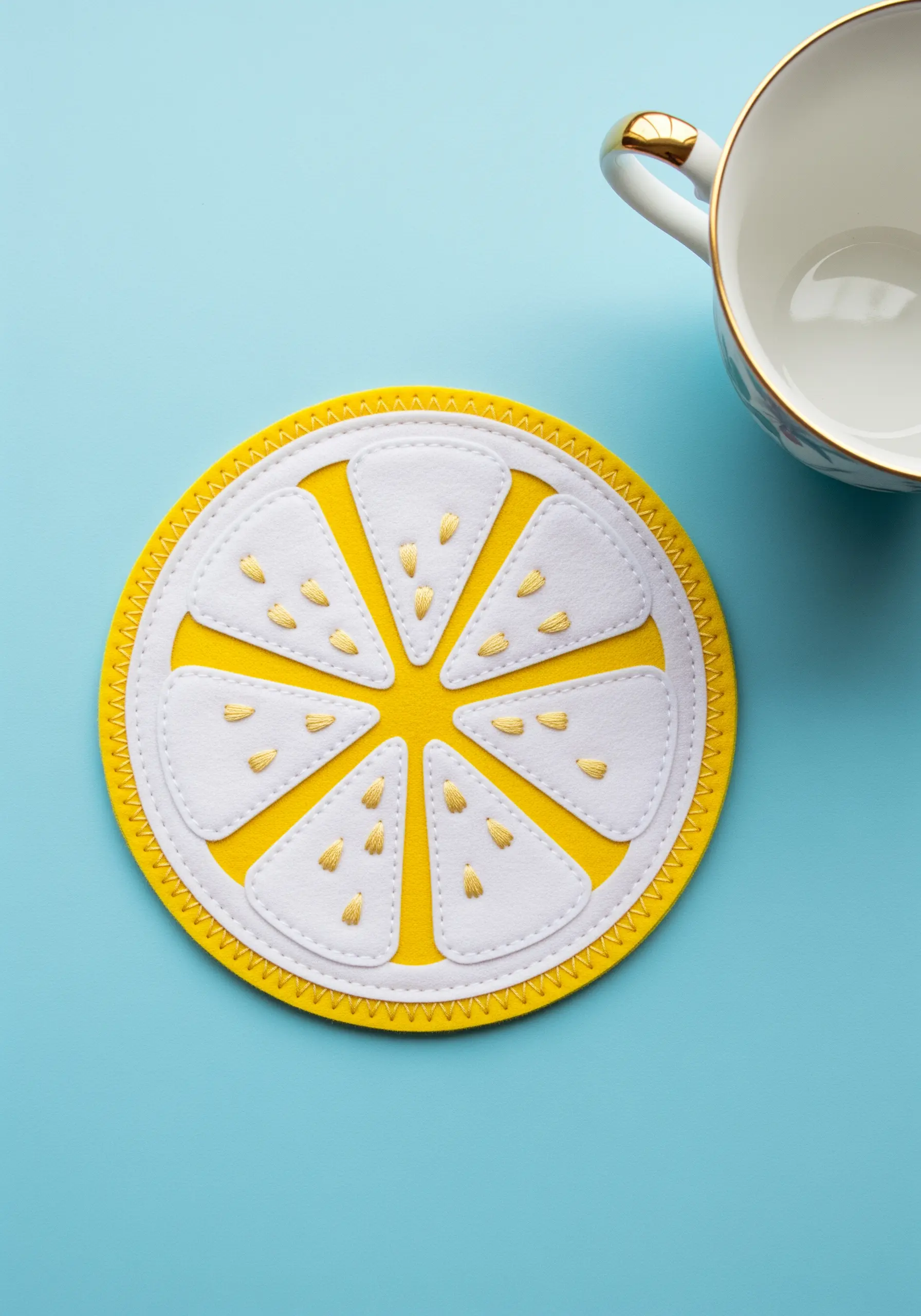 A bright yellow felt coaster shaped like a lemon slice, with white appliqué and gold embroidered seeds.