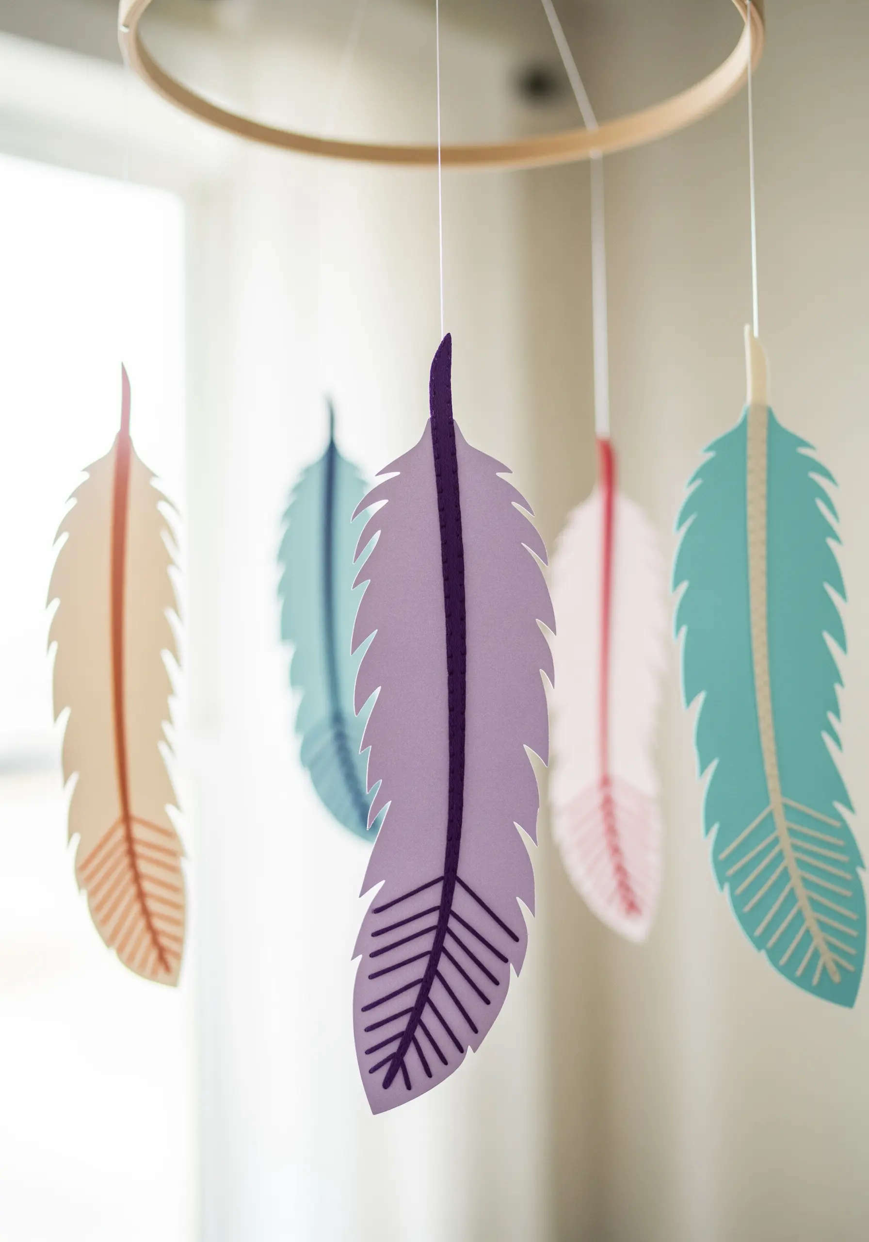 A mobile with five pastel-colored paper feathers, each with stitched details, hanging from a hoop.