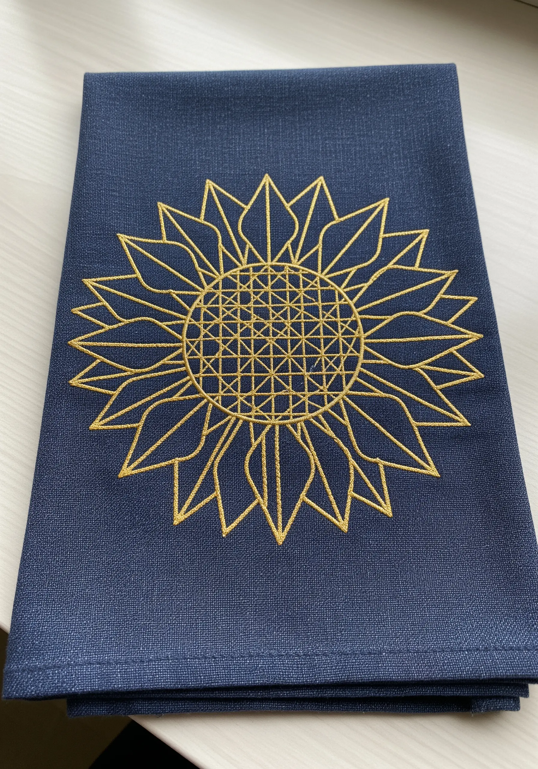 A geometric sunflower embroidered in gold thread on a navy blue towel, showing abstract lines and grids