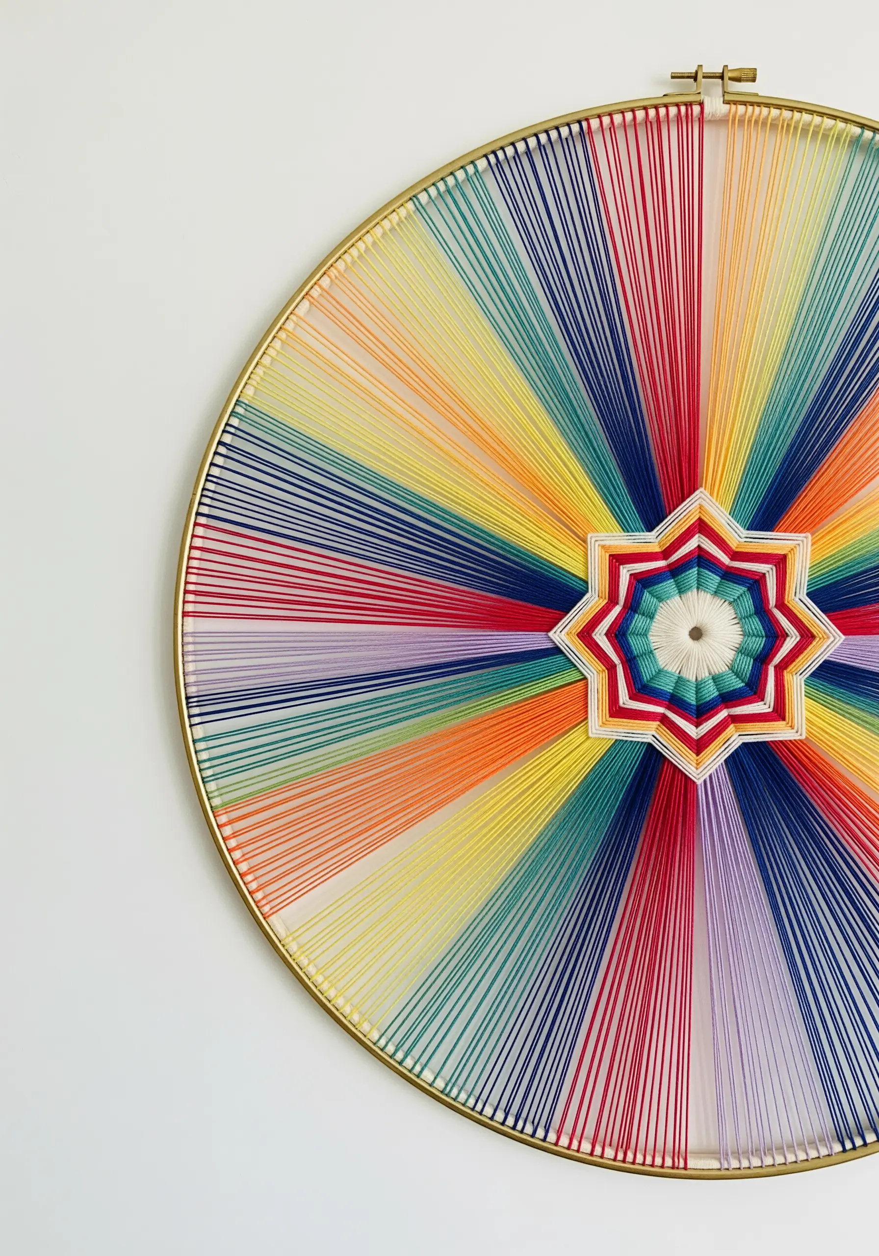 A large metal hoop with colorful threads wrapped to form a rainbow starburst mandala.
