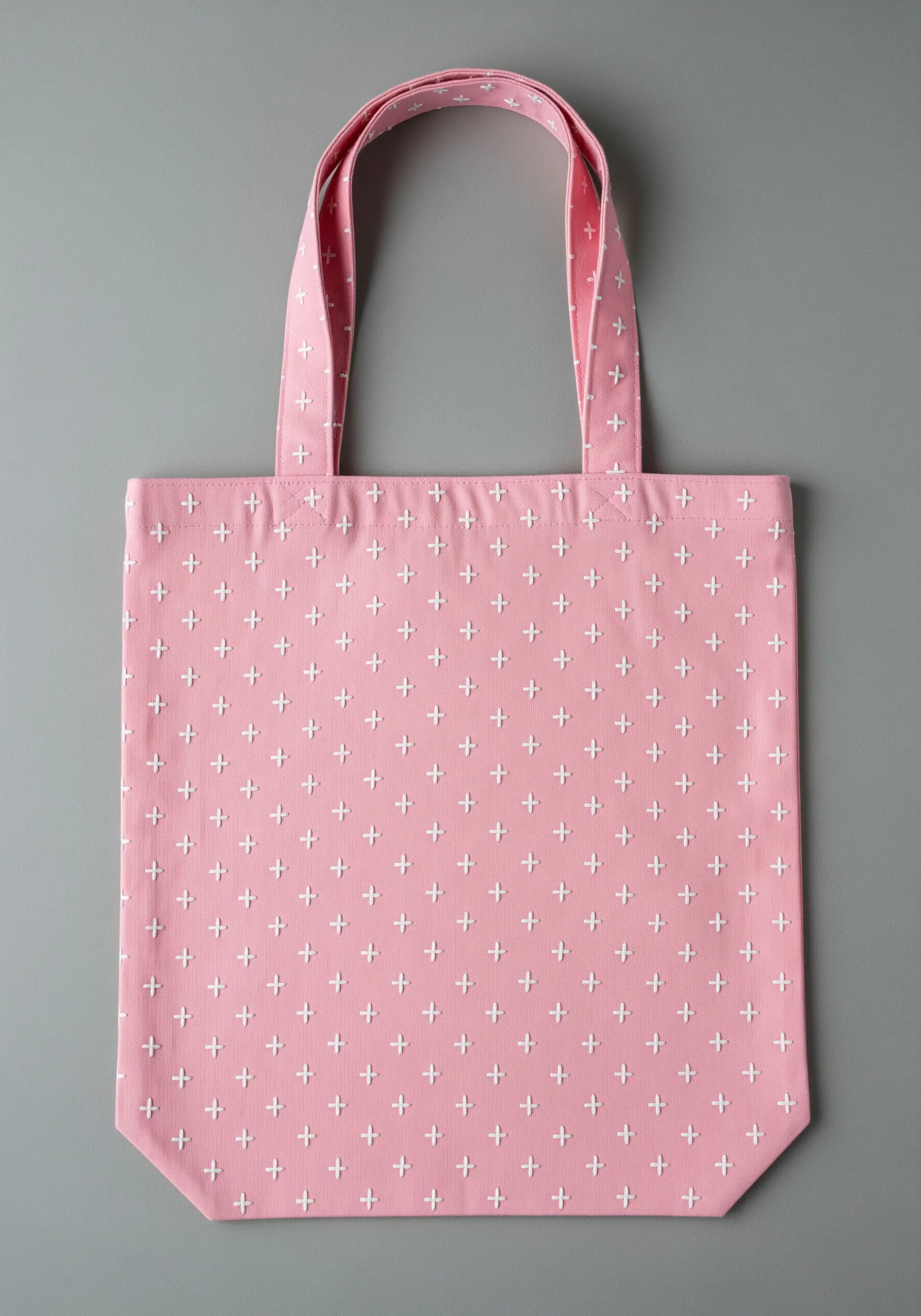 A repeating pattern of small white cross stitches covering a light pink tote bag.