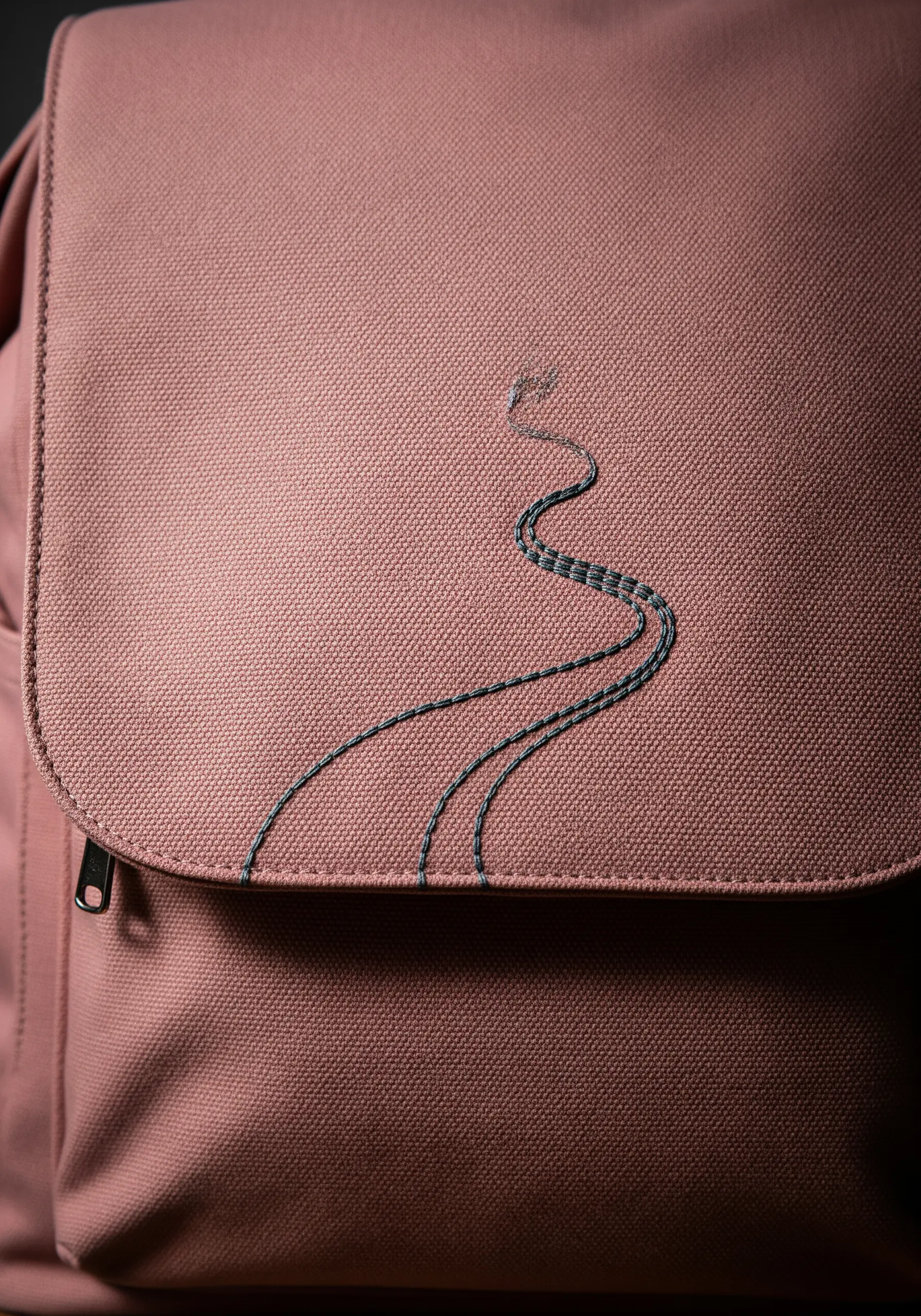 A winding path stitched in variegated thread on the flap of a dusty pink backpack.