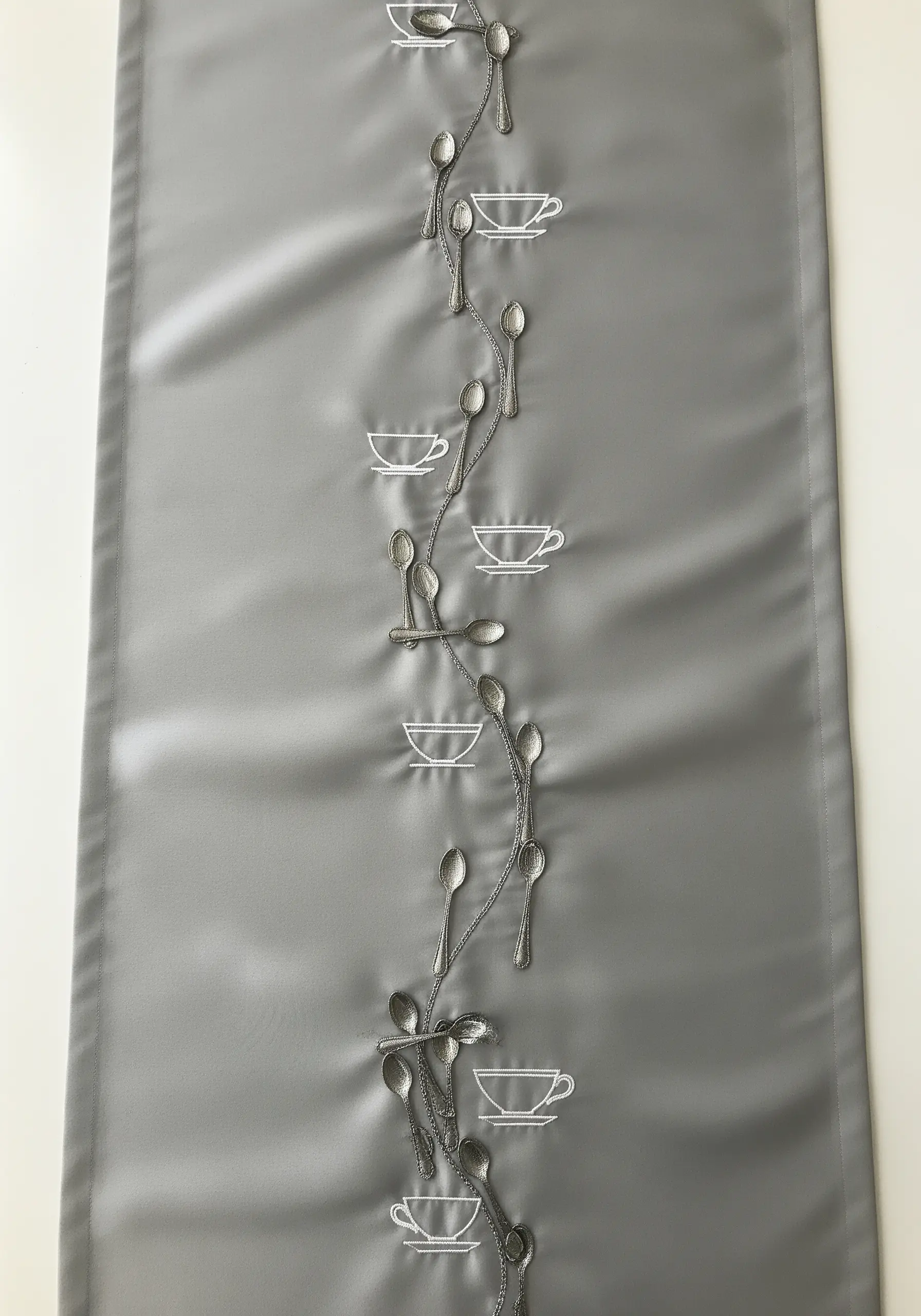 A vertical table runner design with embroidered spoons and teacups connected by a vine.