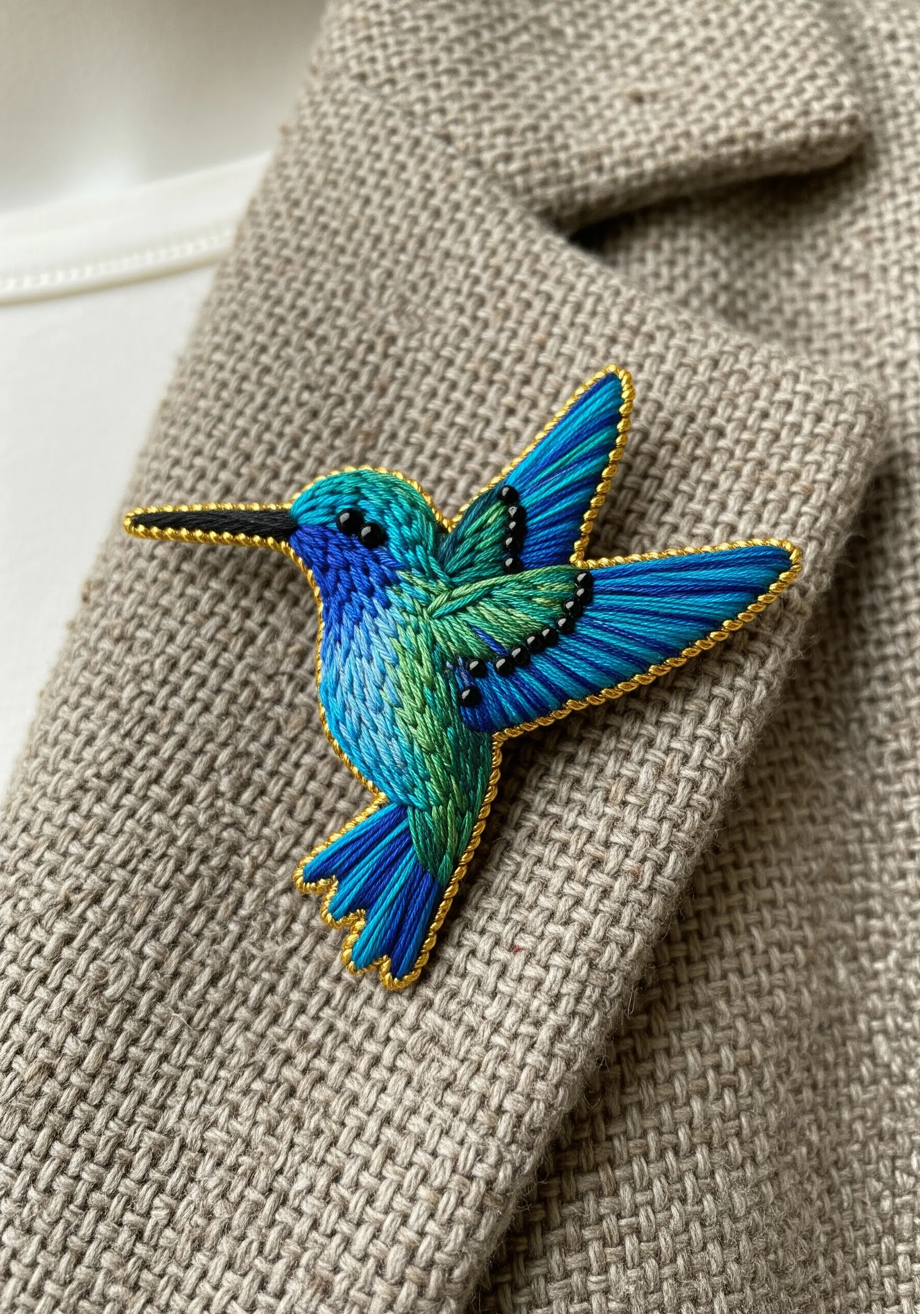 A detailed embroidered hummingbird brooch with thread painting and a clean, couched gold-thread border.