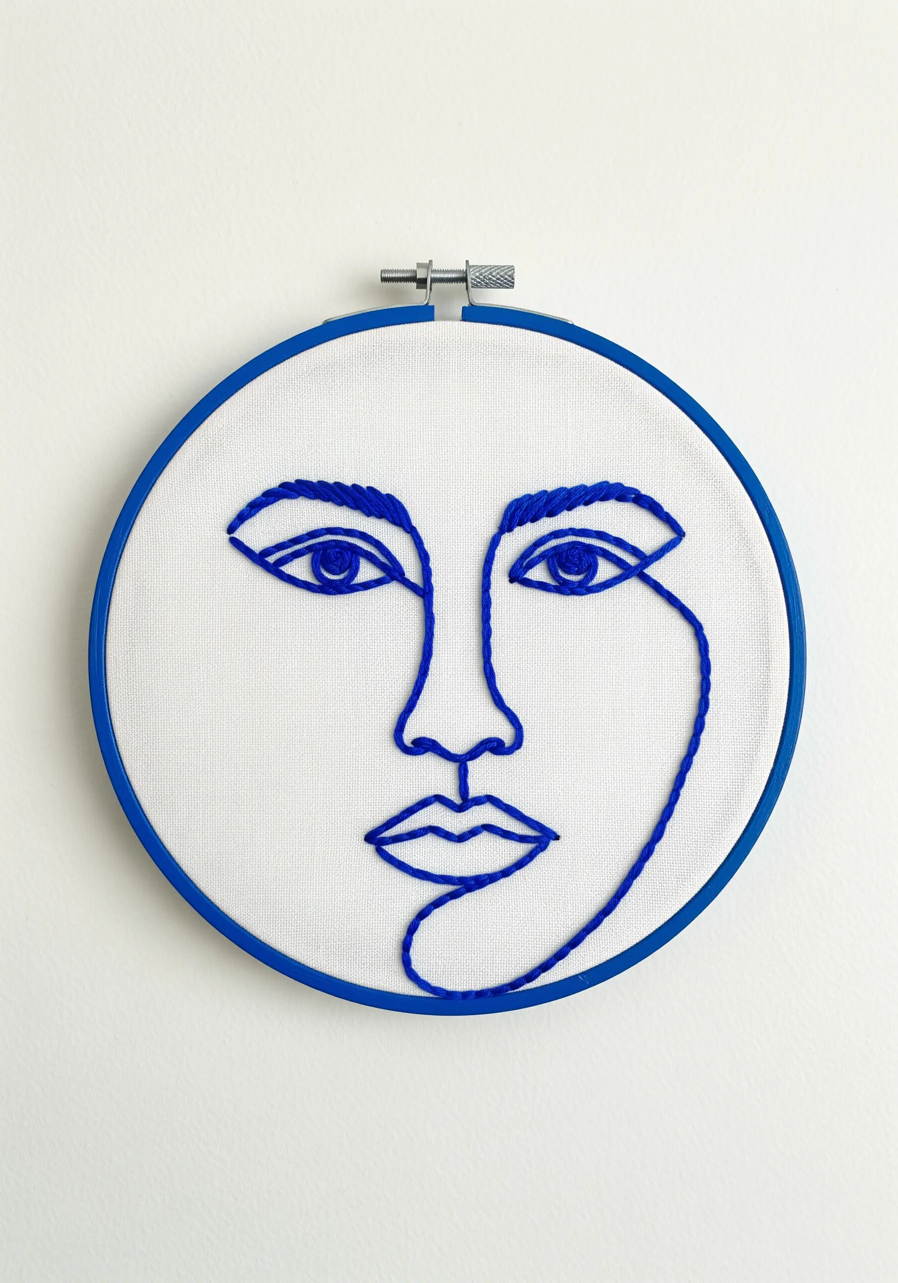 A continuous line face stitched in bold cobalt blue thread on white fabric.