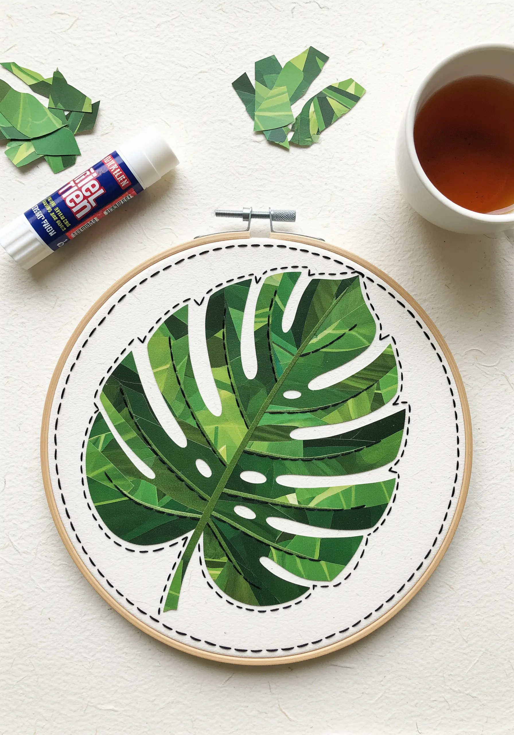 A paper collage monstera leaf outlined with black backstitch on white fabric in a hoop.