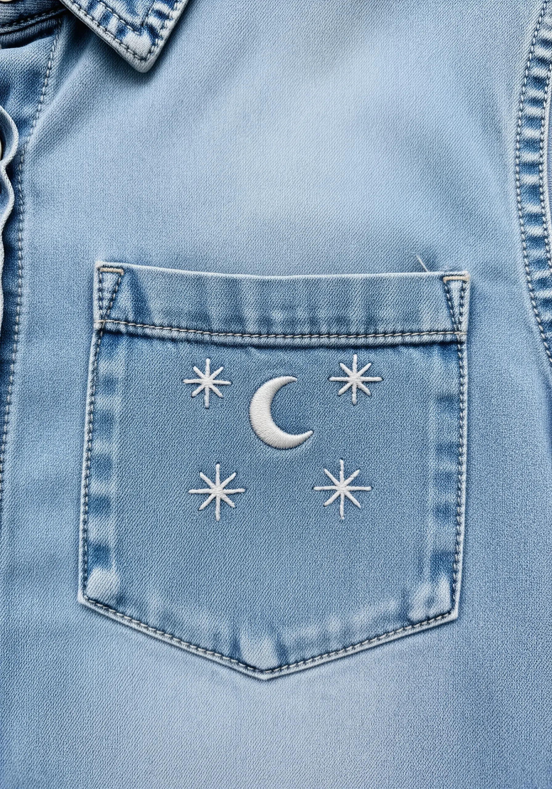 A tiny crescent moon and stars embroidered in white thread on a denim shirt pocket.
