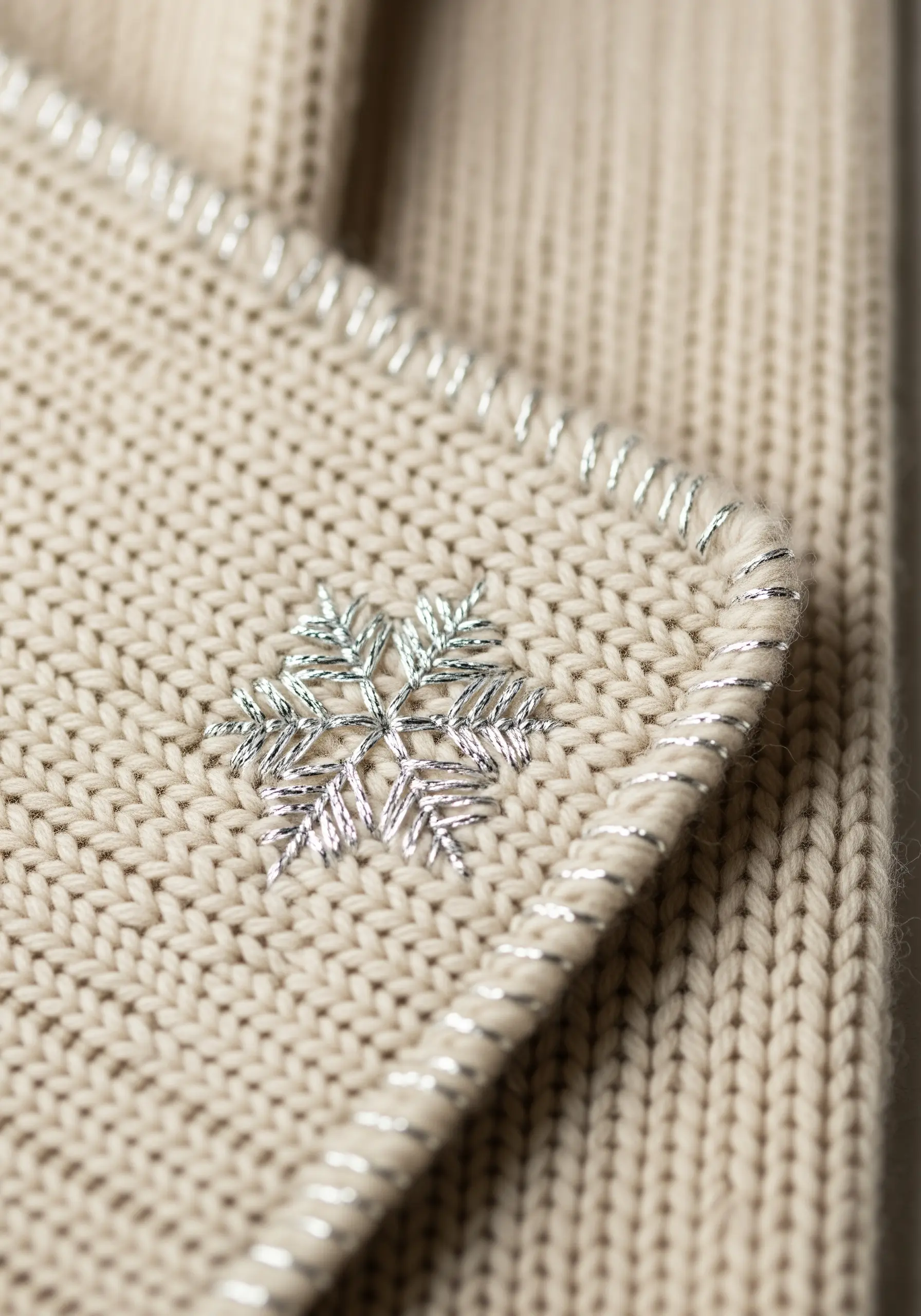 A delicate silver metallic snowflake embroidered on the corner of a cream-colored knit scarf.