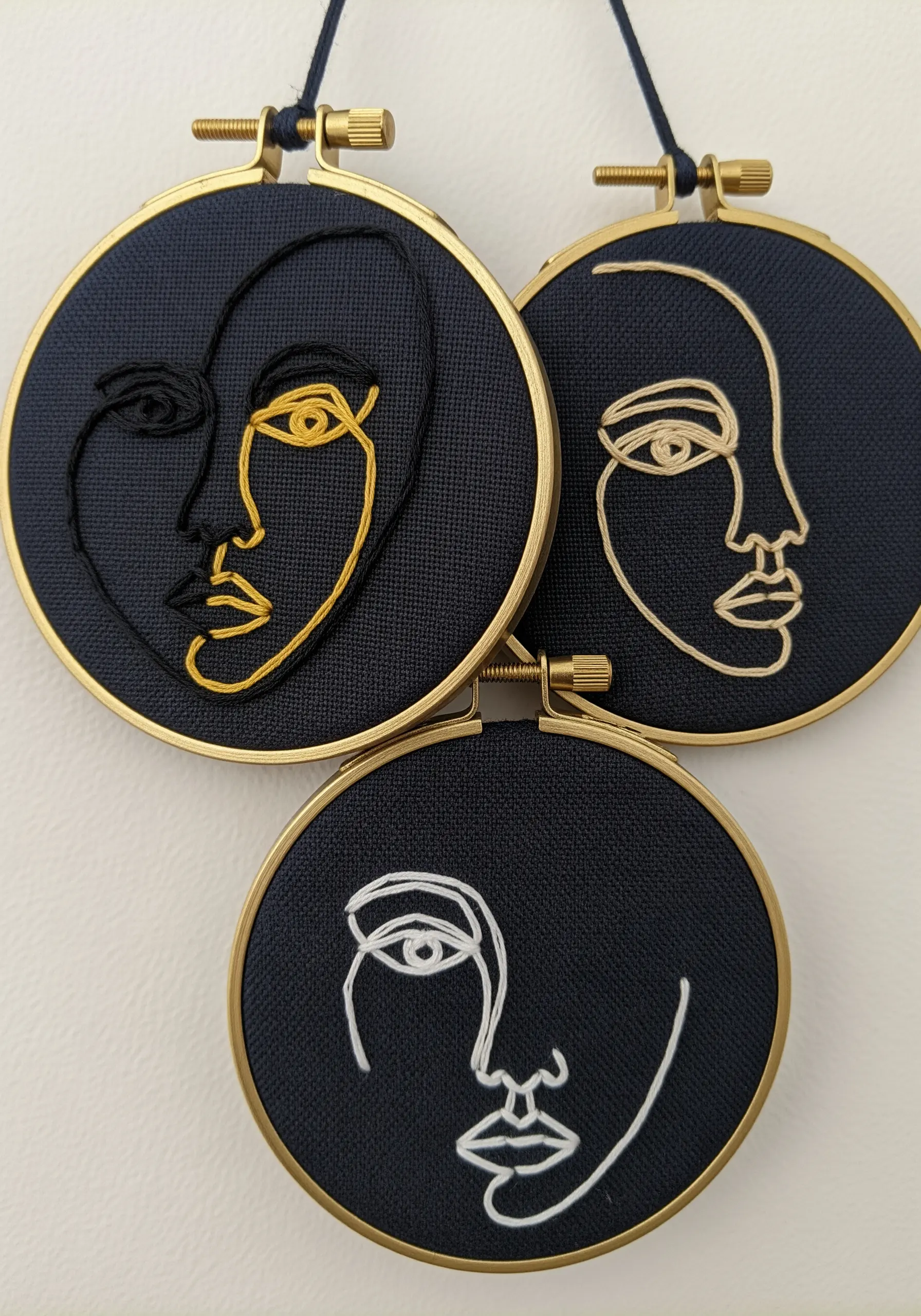 Three small hoops on dark navy fabric, each with a line art face in black, white, or gold.