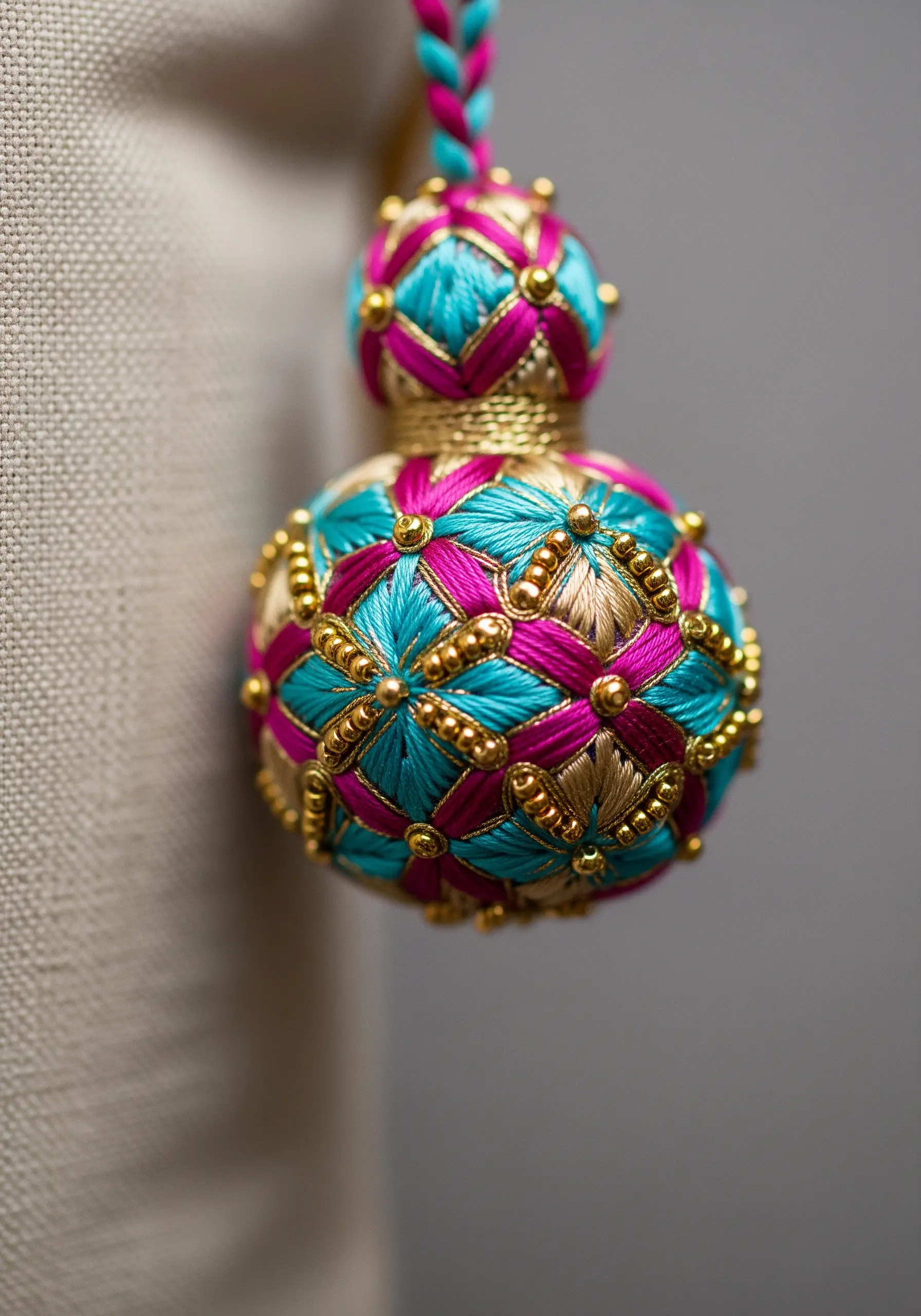 An ornate, decorative tassel made from wrapped turquoise, pink, and gold thread with beaded accents.