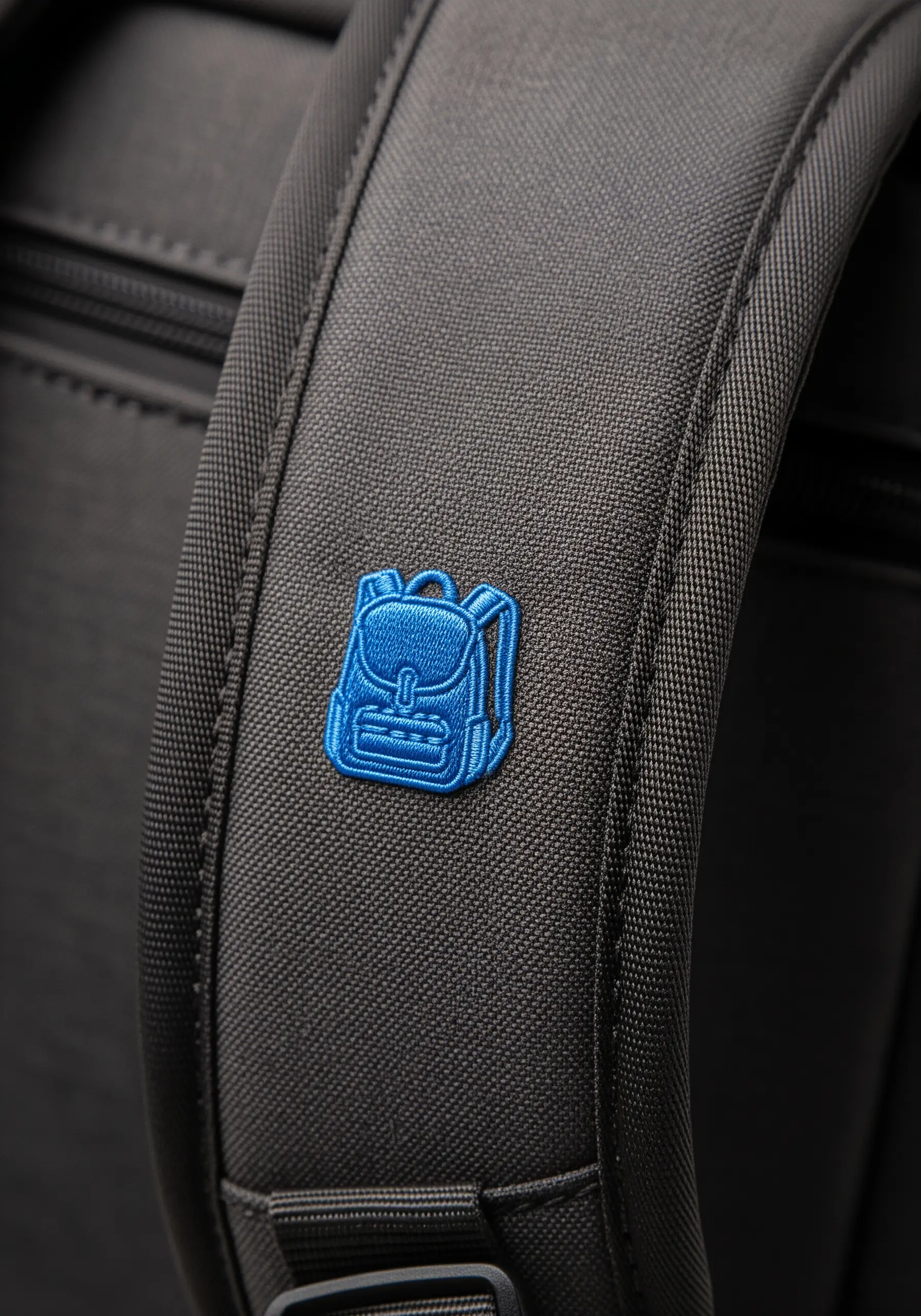 A tiny, bright blue backpack icon embroidered on the strap of a larger black backpack.