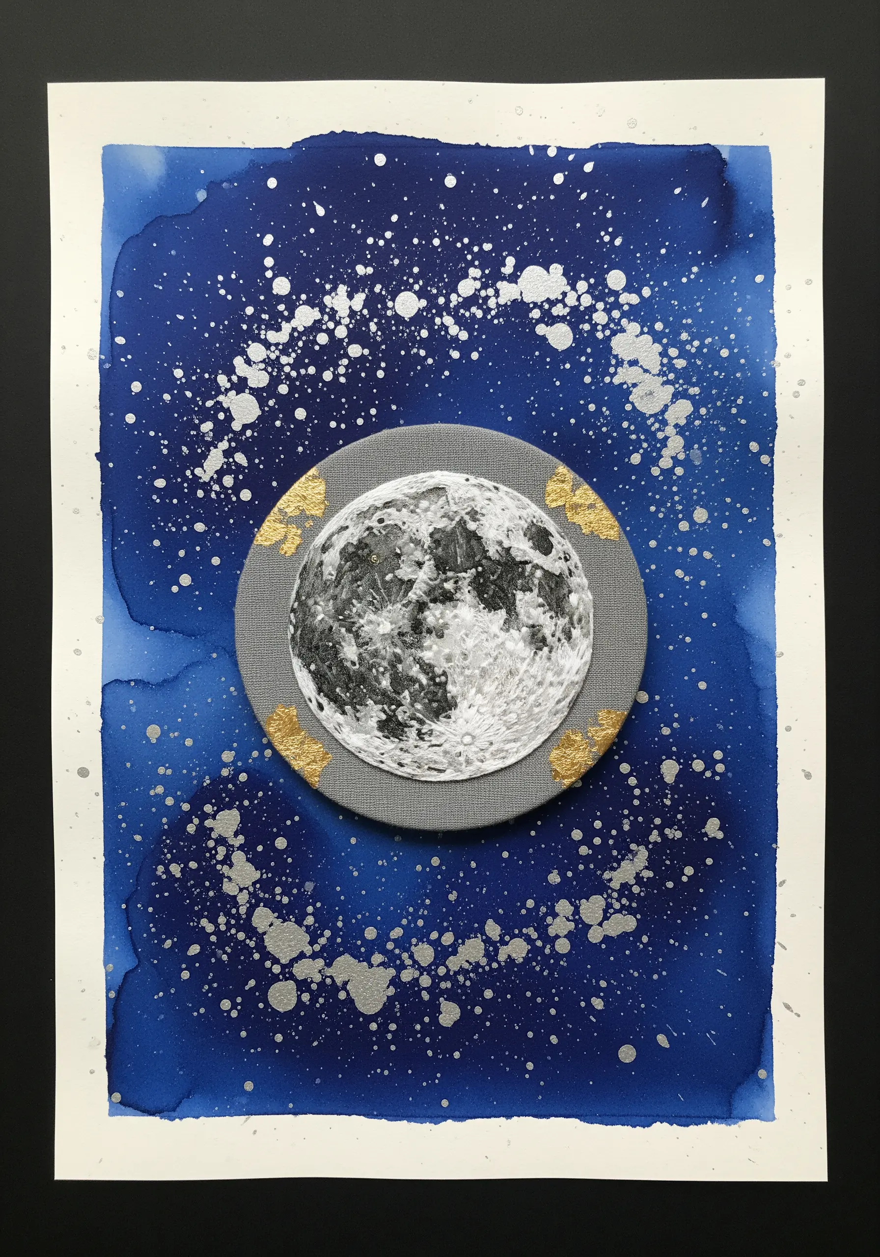 An embroidered moon with gold leaf details, mounted on a dark blue and silver splattered watercolor paper.