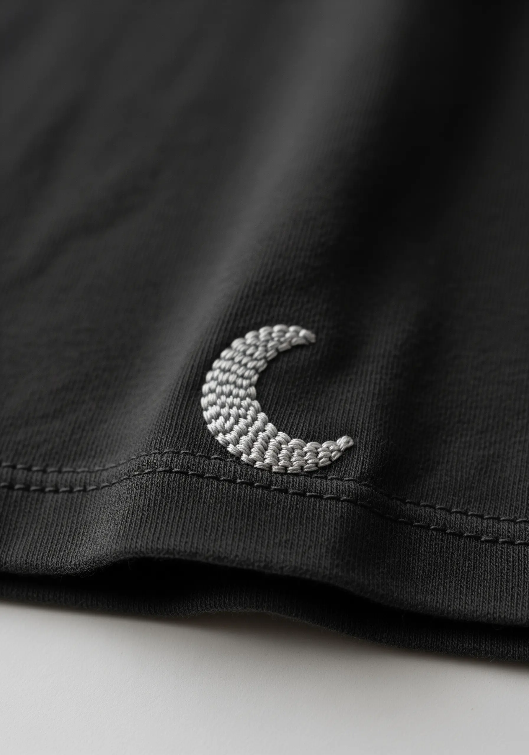 A textured, light grey embroidered crescent moon on a dark grey t-shirt.