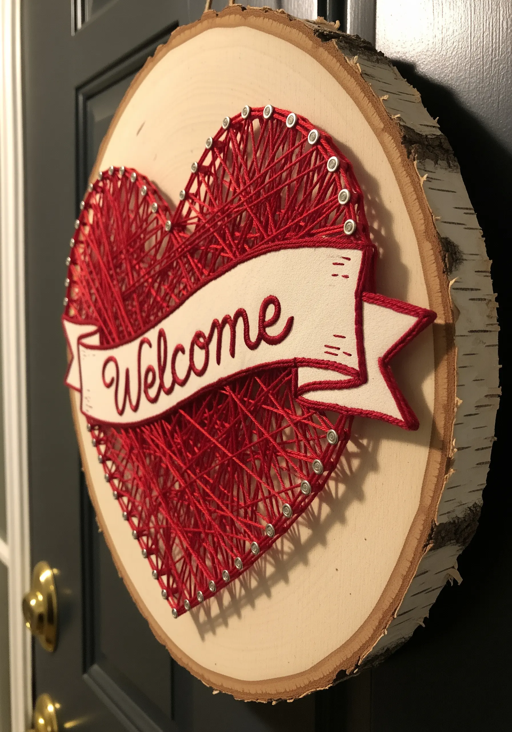 A red string art heart on a wood slice with an embroidered 'Welcome' banner layered on top.
