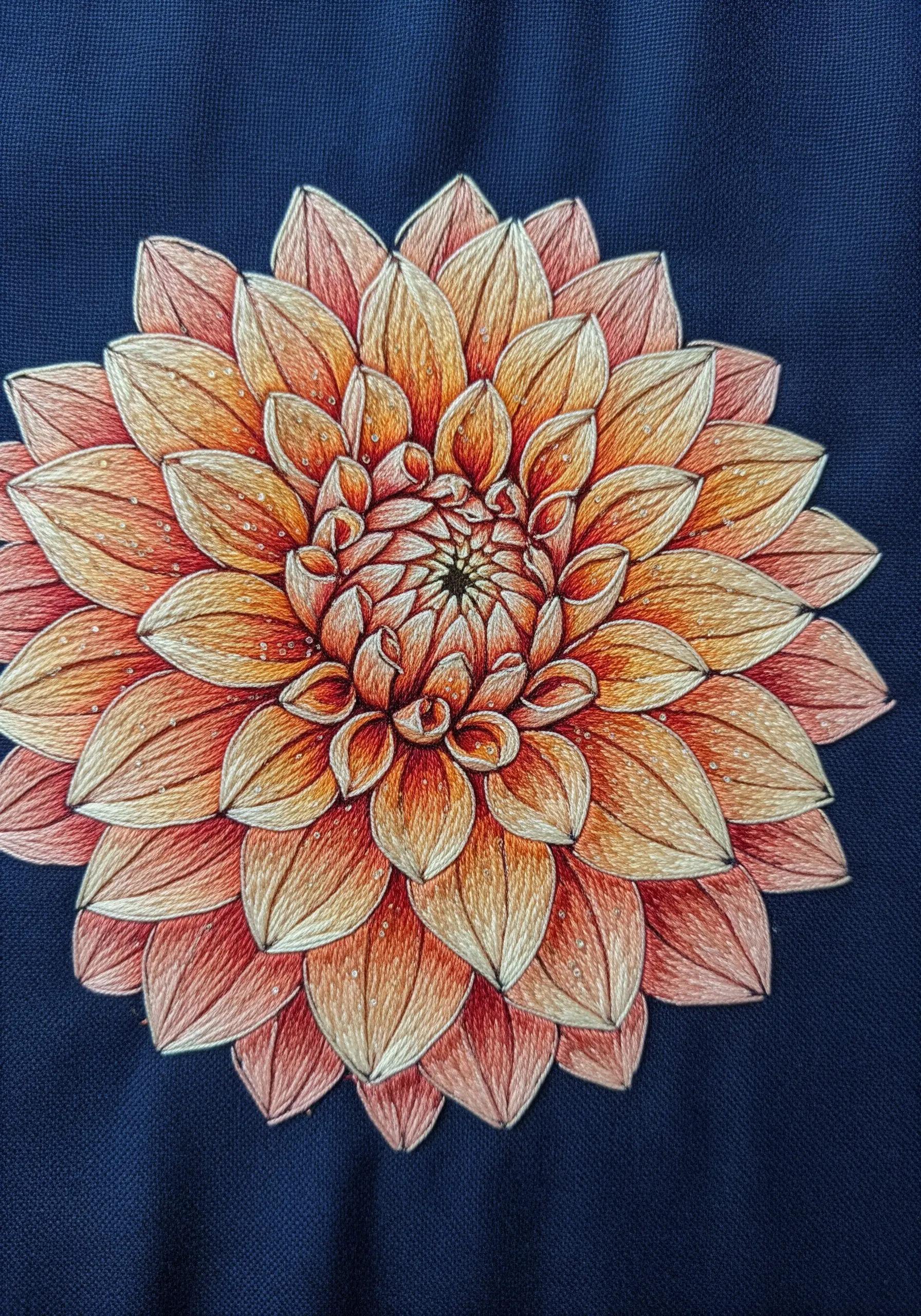 A thread-painted dahlia with orange and coral petals on a dark blue background.