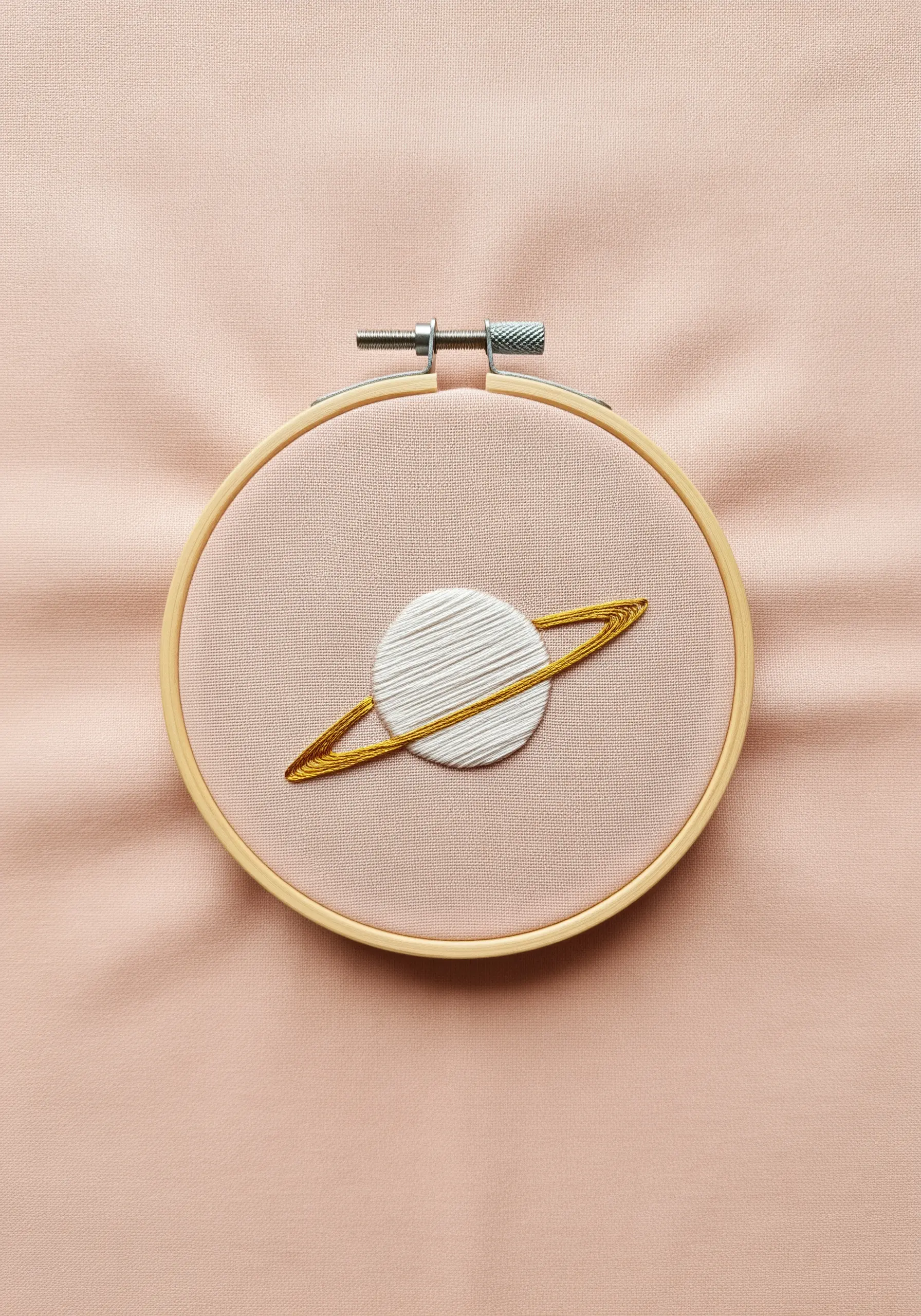 A planet with white satin stitches and gold metallic rings on pale pink fabric.