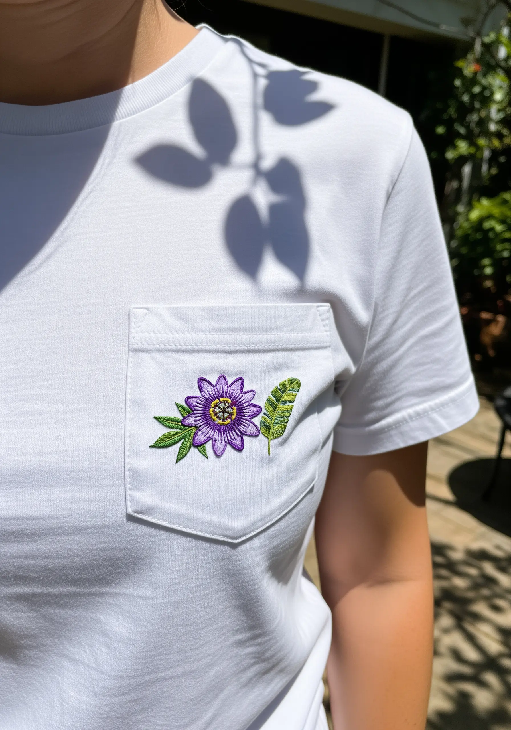 A detailed passion flower and a small leaf embroidered on the pocket of a white t-shirt.