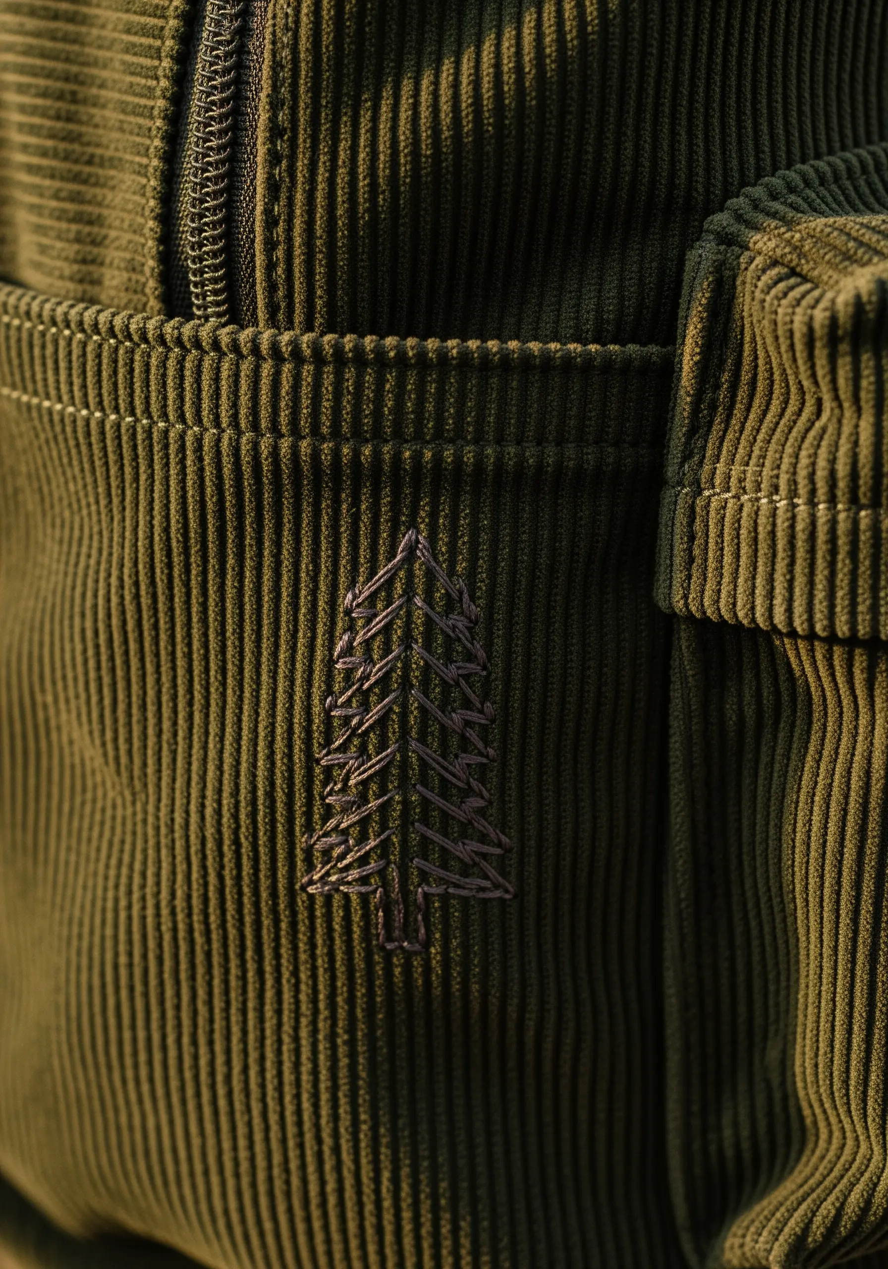 A simple line-art pine tree embroidered in dark thread on the pocket of an olive corduroy backpack.