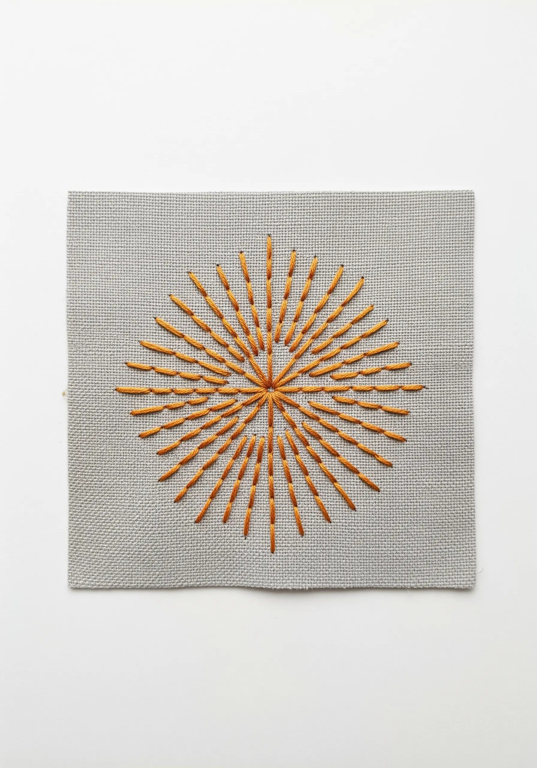 A radiating sunburst motif in golden-orange thread on gray Aida cloth.