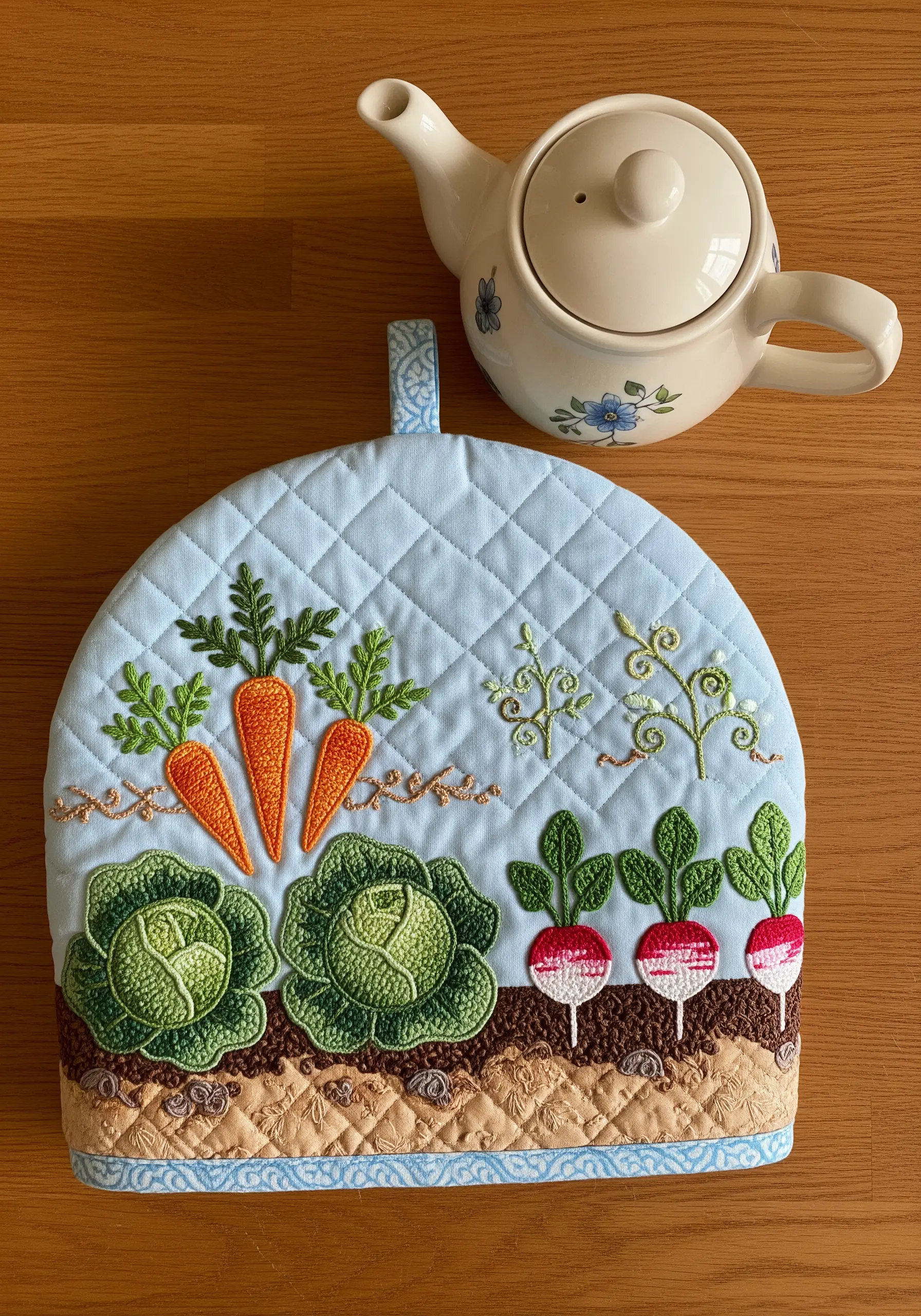 A quilted tea cozy decorated with appliqué and embroidered vegetables like carrots and cabbage.