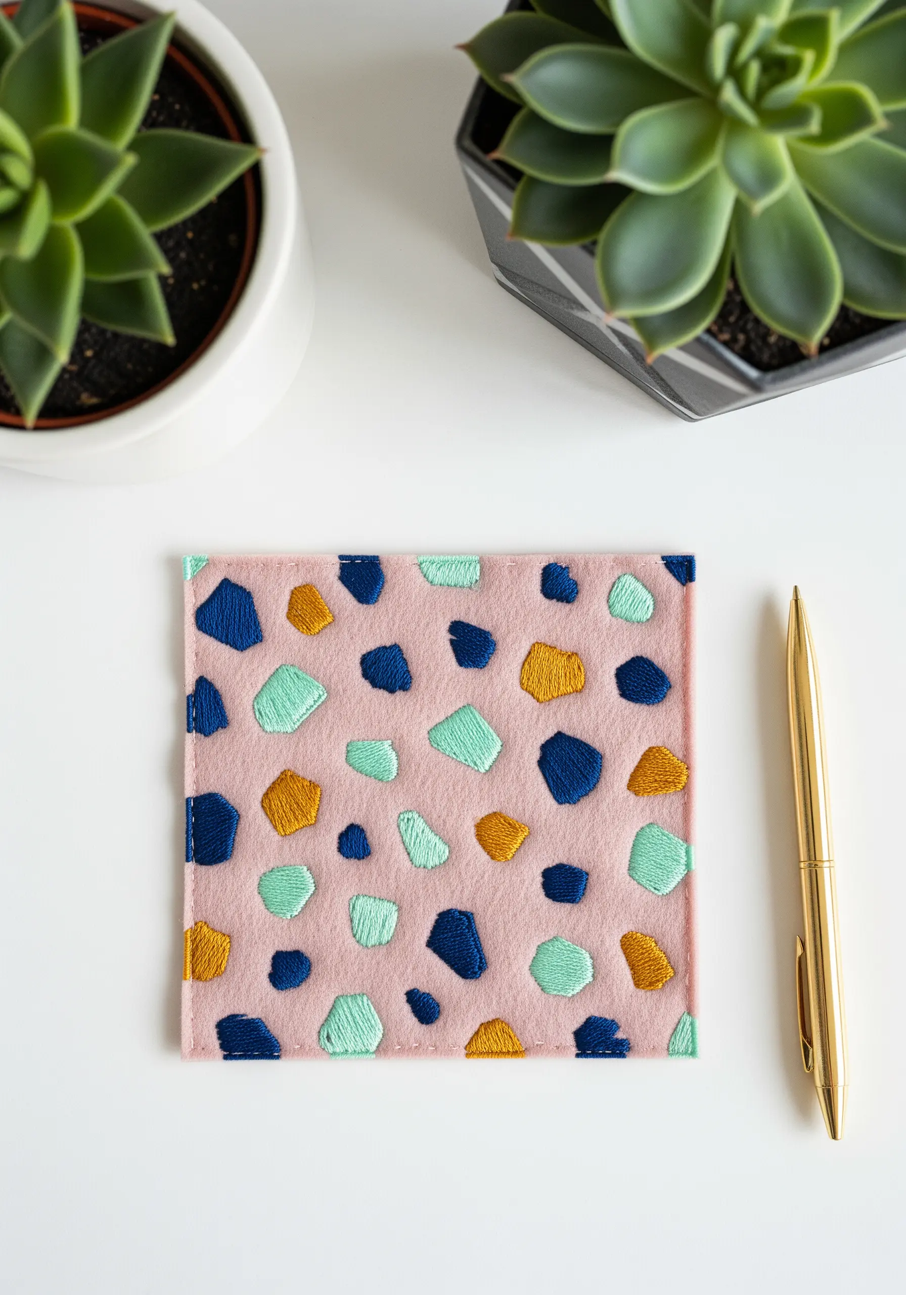 A modern terrazzo pattern embroidered with satin-stitched shapes in blue, gold, and mint on pink fabric.