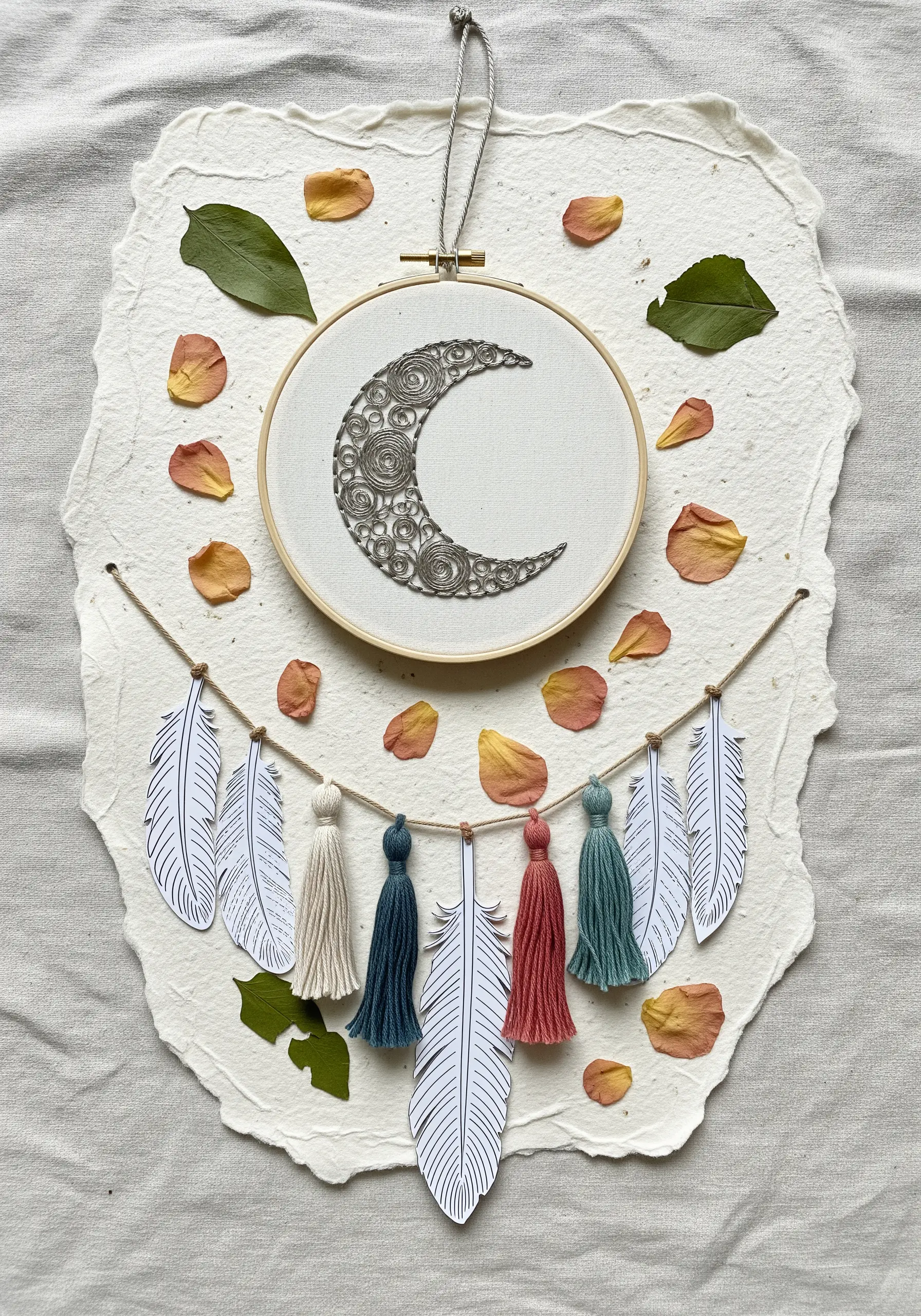 An embroidered moon in a hoop, mounted on a large sheet of handmade paper with dried petals.