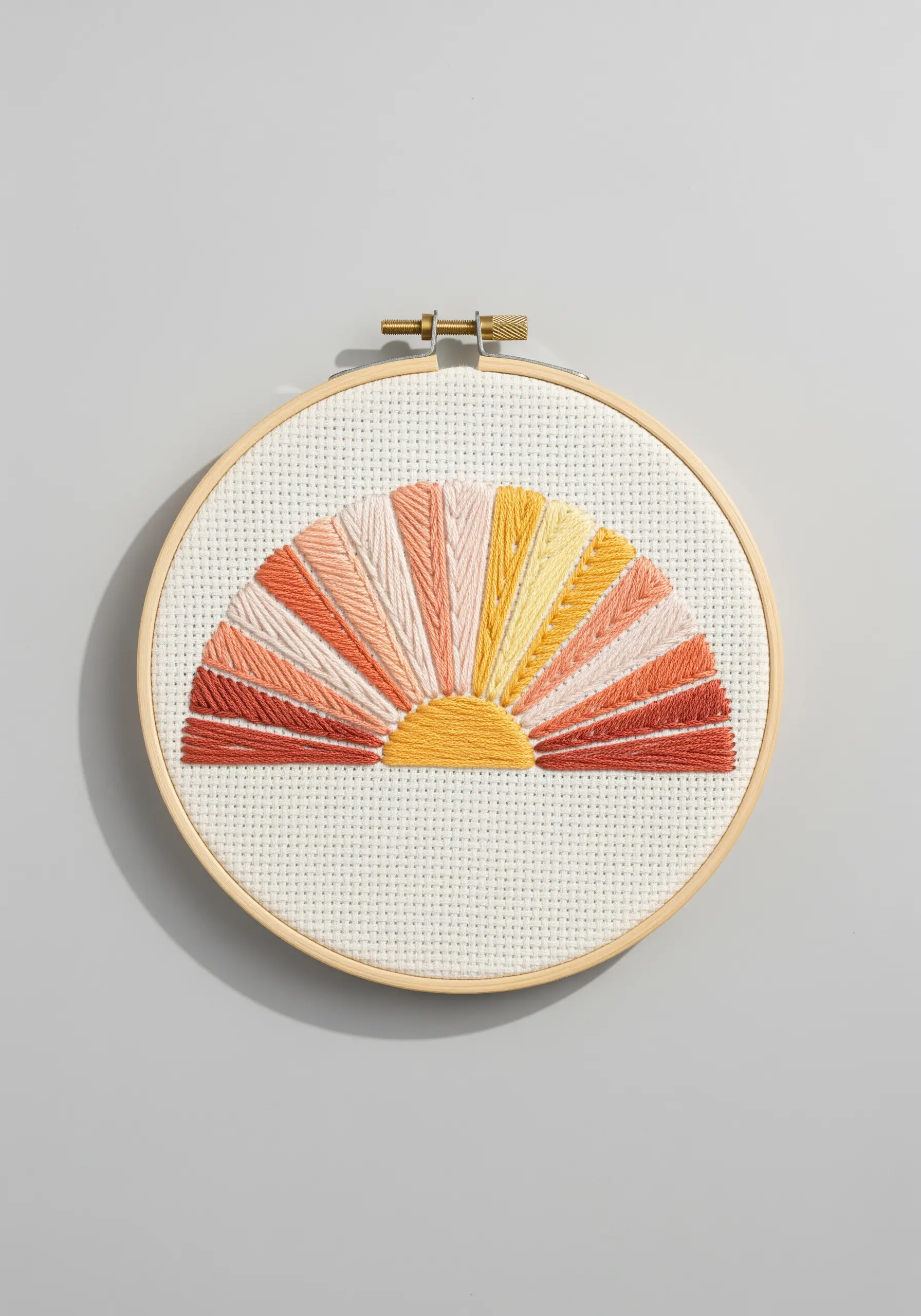 Abstract sunrise embroidery with radiating rays in warm tones on Aida cloth.