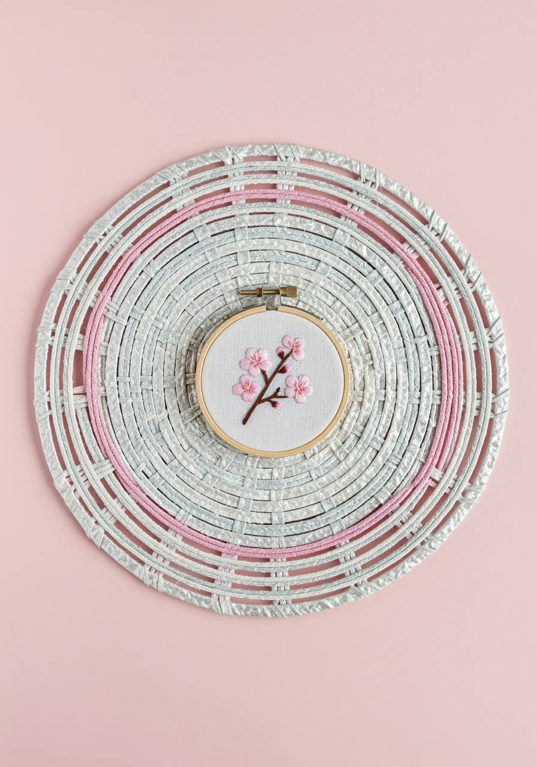 A small embroidery hoop with pink cherry blossoms, centered on a woven silver and pink paper placemat.