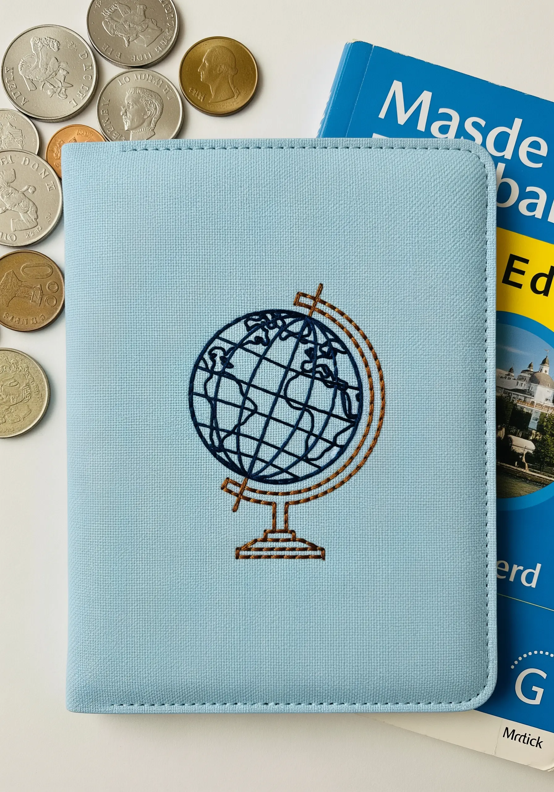 A line-art globe embroidered in blue and gold thread on a light blue passport cover.
