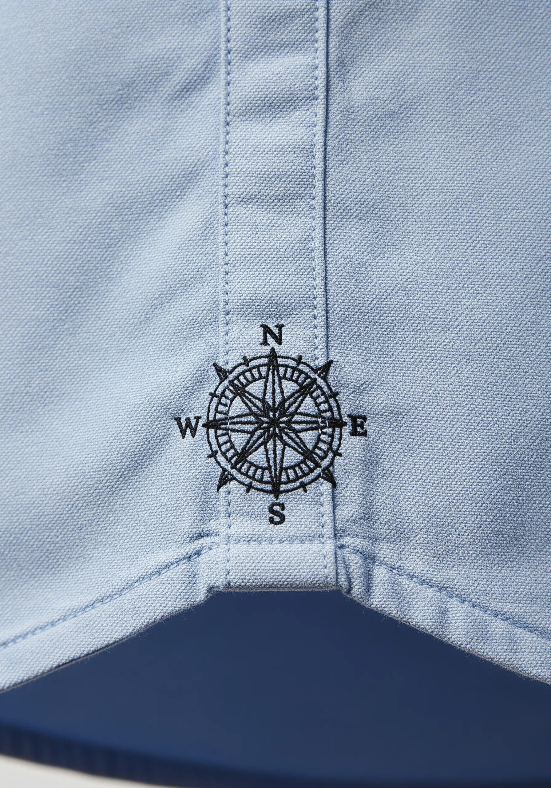 A black compass rose with cardinal directions embroidered on the front placket of a light blue shirt.