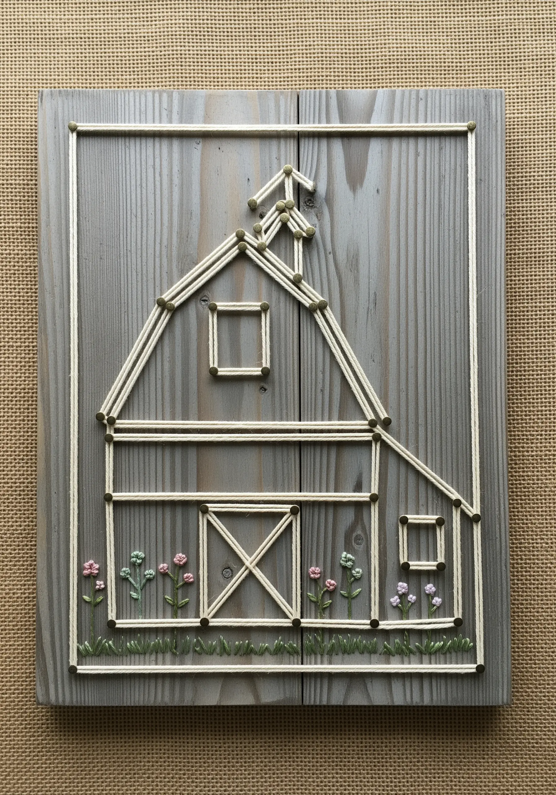 String art barn on a grey wood panel with small embroidered flowers at its base.