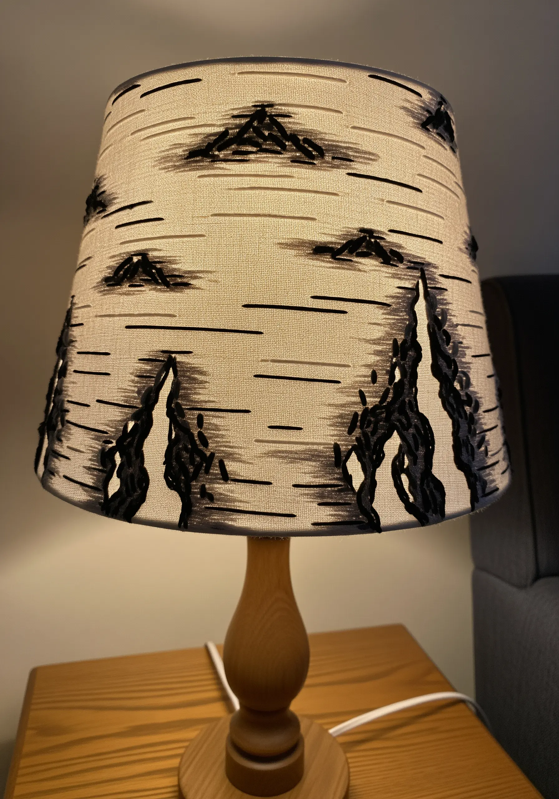 A fabric lampshade embroidered with black stitches to look like birch bark, lit from within.