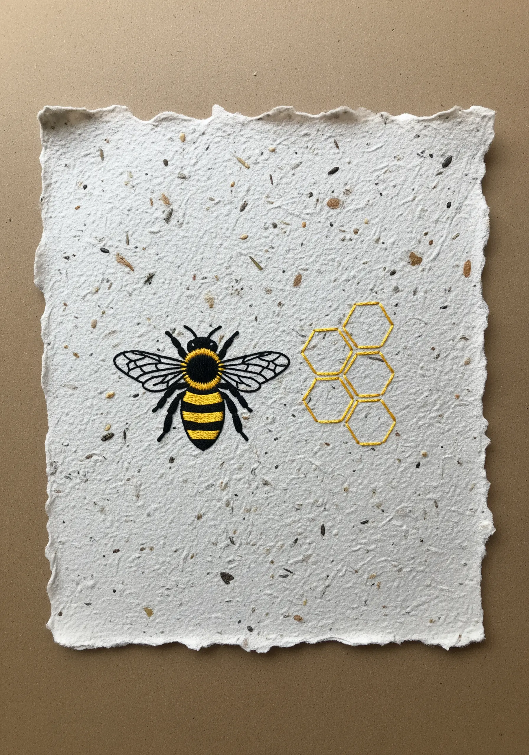 A neatly embroidered bee and honeycomb outline on a piece of handmade paper with visible seeds.