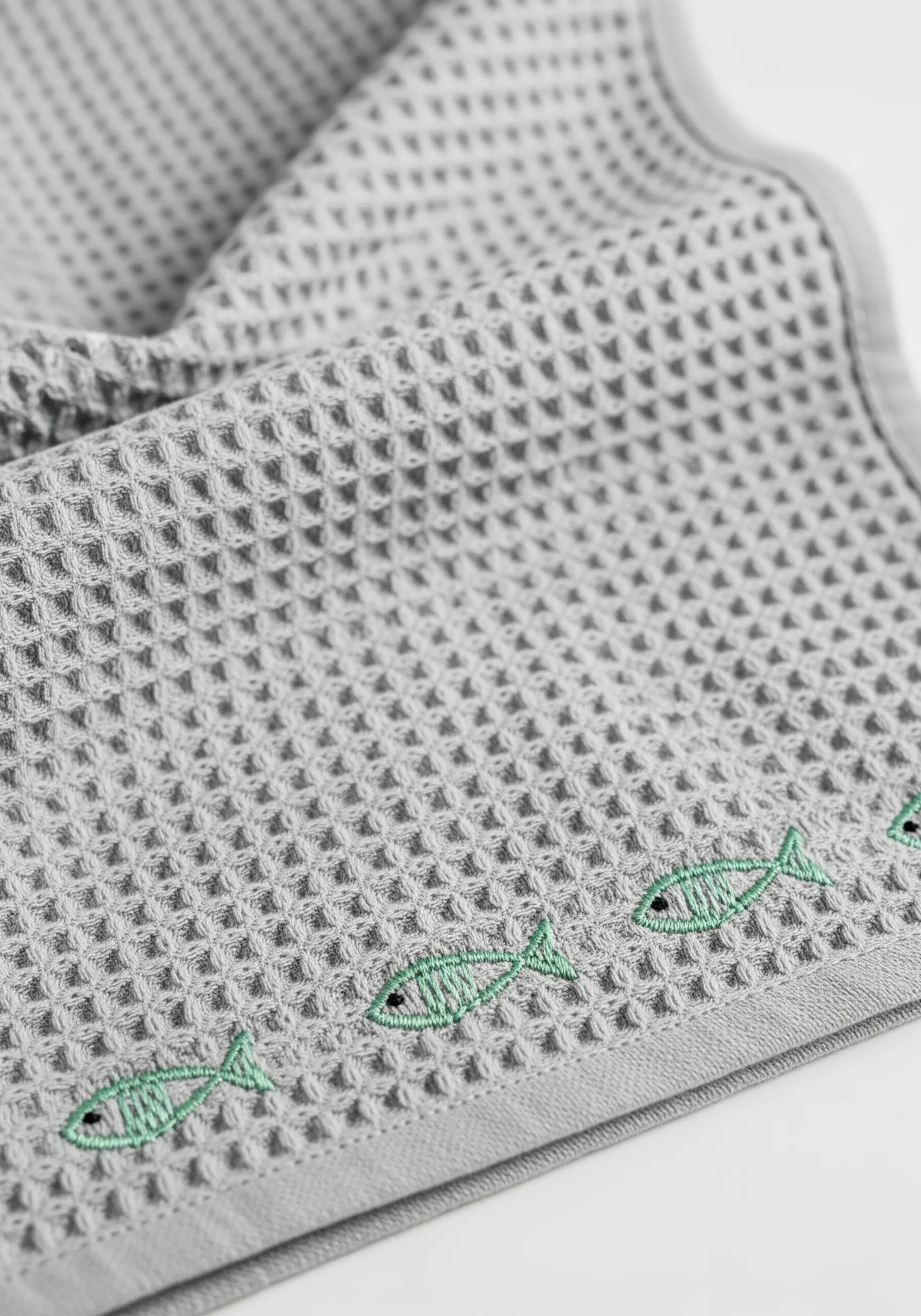 A border of small green fish embroidered on a grey waffle-weave kitchen towel.