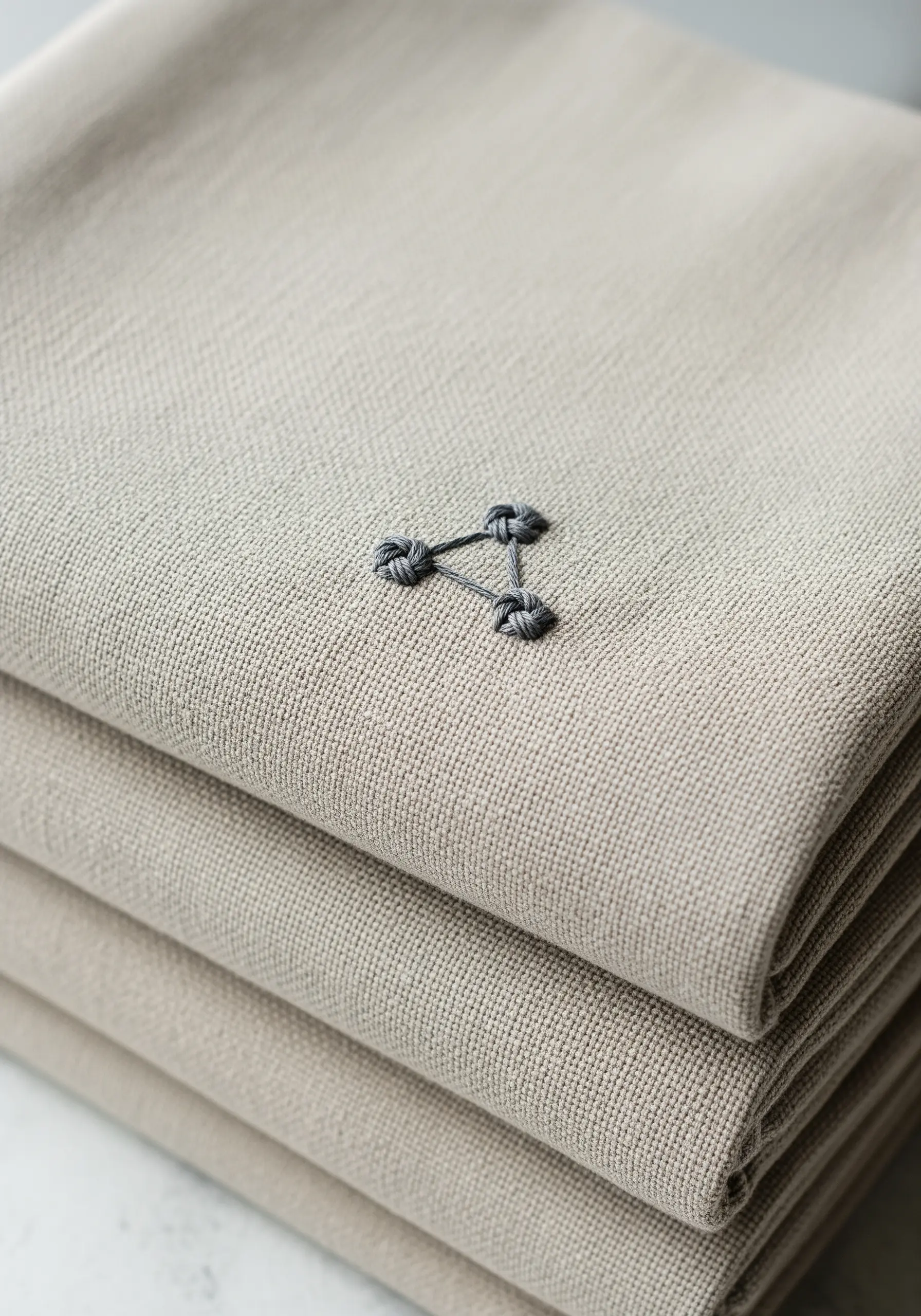 A small, textured triangular knot embroidered in grey thread on linen.