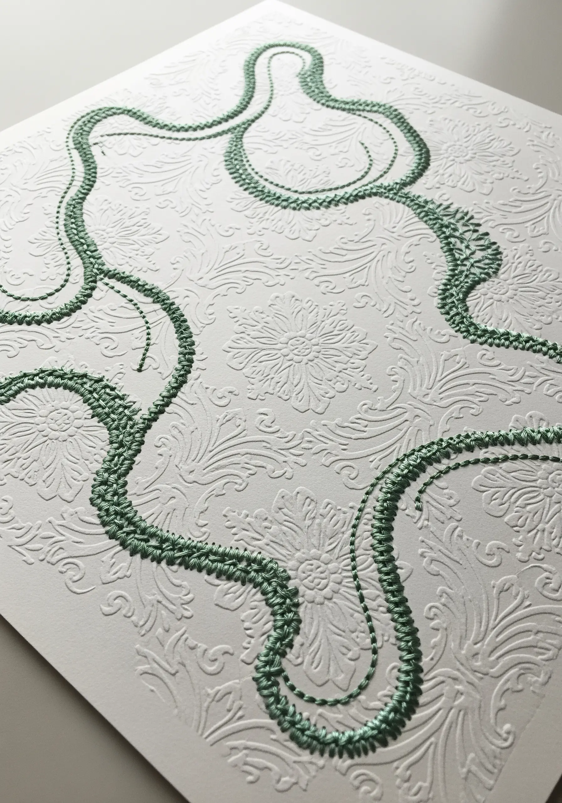 A green chain stitch follows the curves of a floral pattern on white embossed paper.