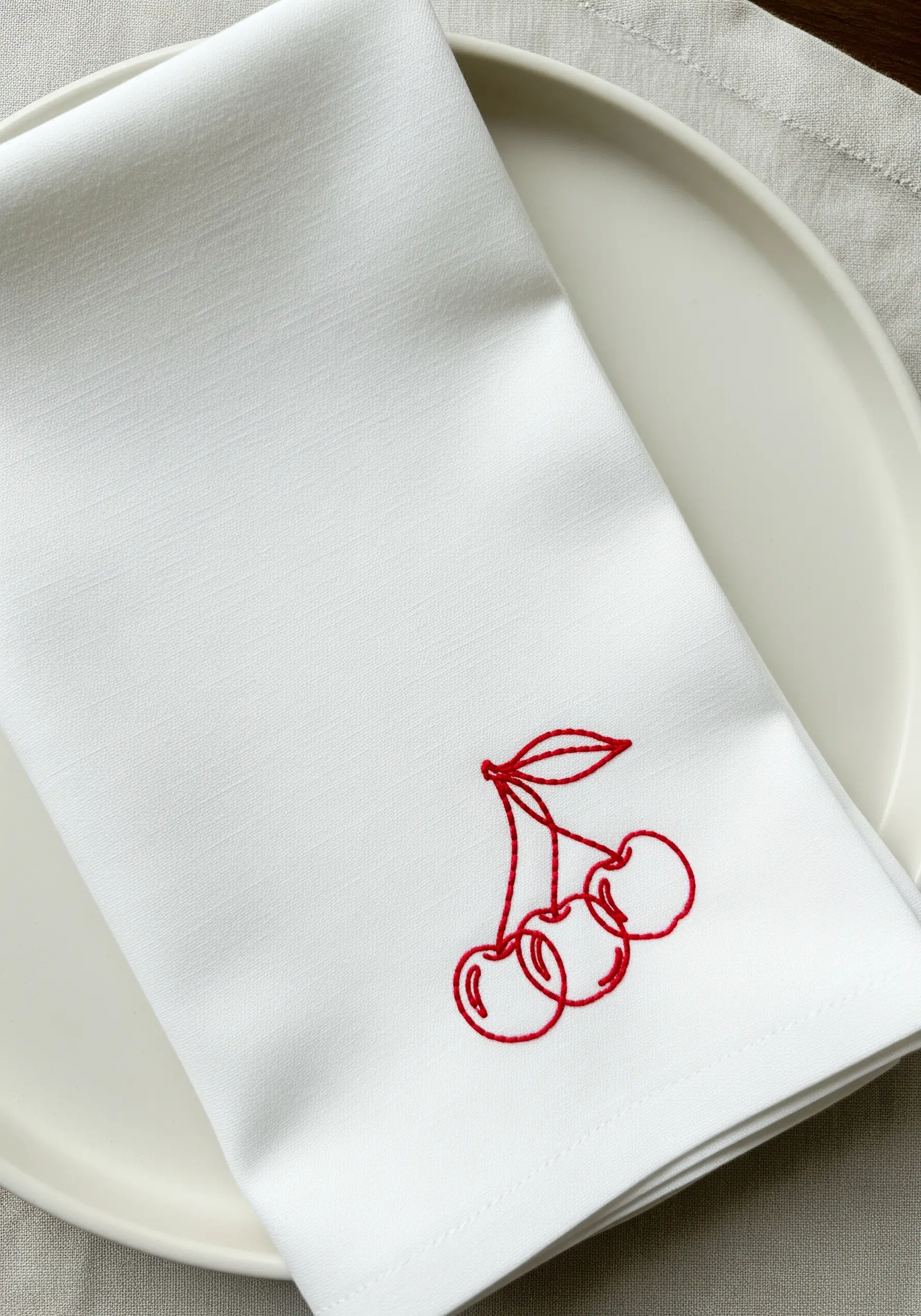 Minimalist red line-art embroidery of three cherries on a white napkin.